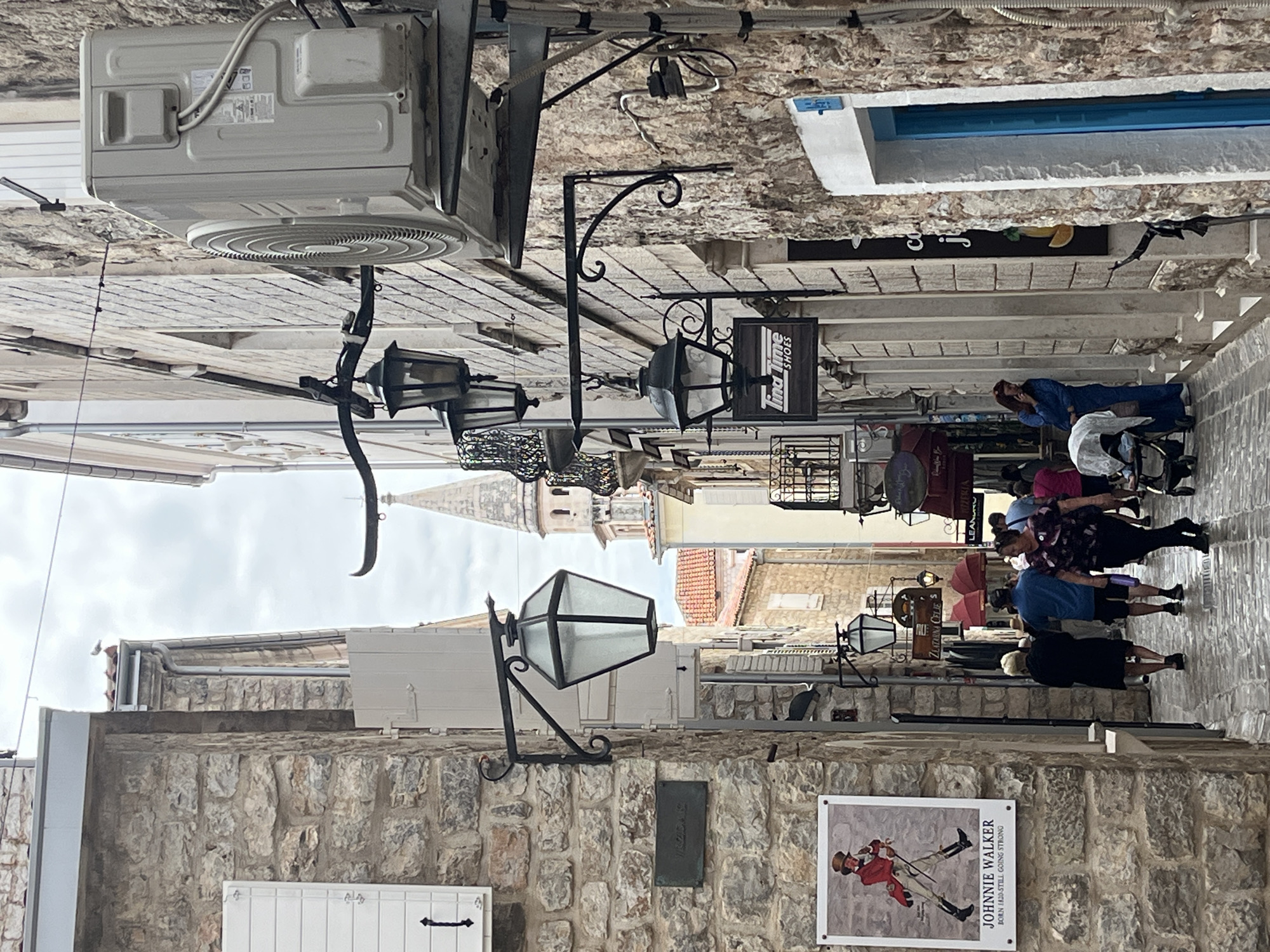 The image depicts a narrow street in an urban area. The street is lined with buildings constructed of stone. An air conditioning unit is mounted high on one of the buildings. Period lamps are affixed to the stone walls. People are walking along the street. Signs for shops are also visible. A sign for "Johnnie Walker" is affixed to a wall. A church steeple is visible in the distance.