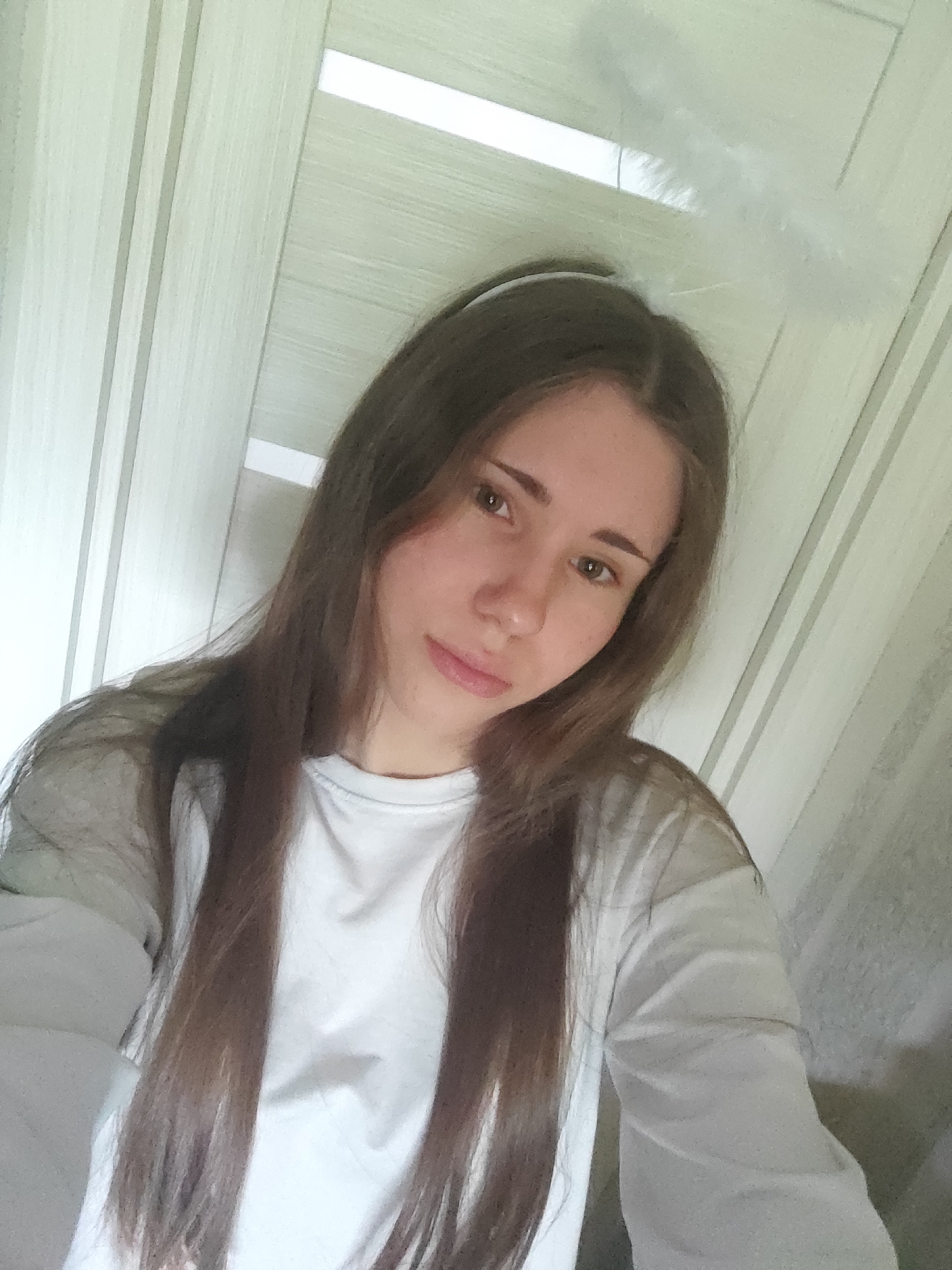 A young woman with long, light brown hair is taking a selfie in an indoor setting. She is wearing a white long-sleeved shirt. A thin white headband is visible on her head, with a fluffy white feather-like object extending from it, suggesting it is an accessory, possibly a halo. The background consists of a light-colored wooden door with vertical paneling and thin white horizontal accents. The lighting is soft and natural. The image appears to be taken in a residential interior in Svietlahorsk, Belarus.