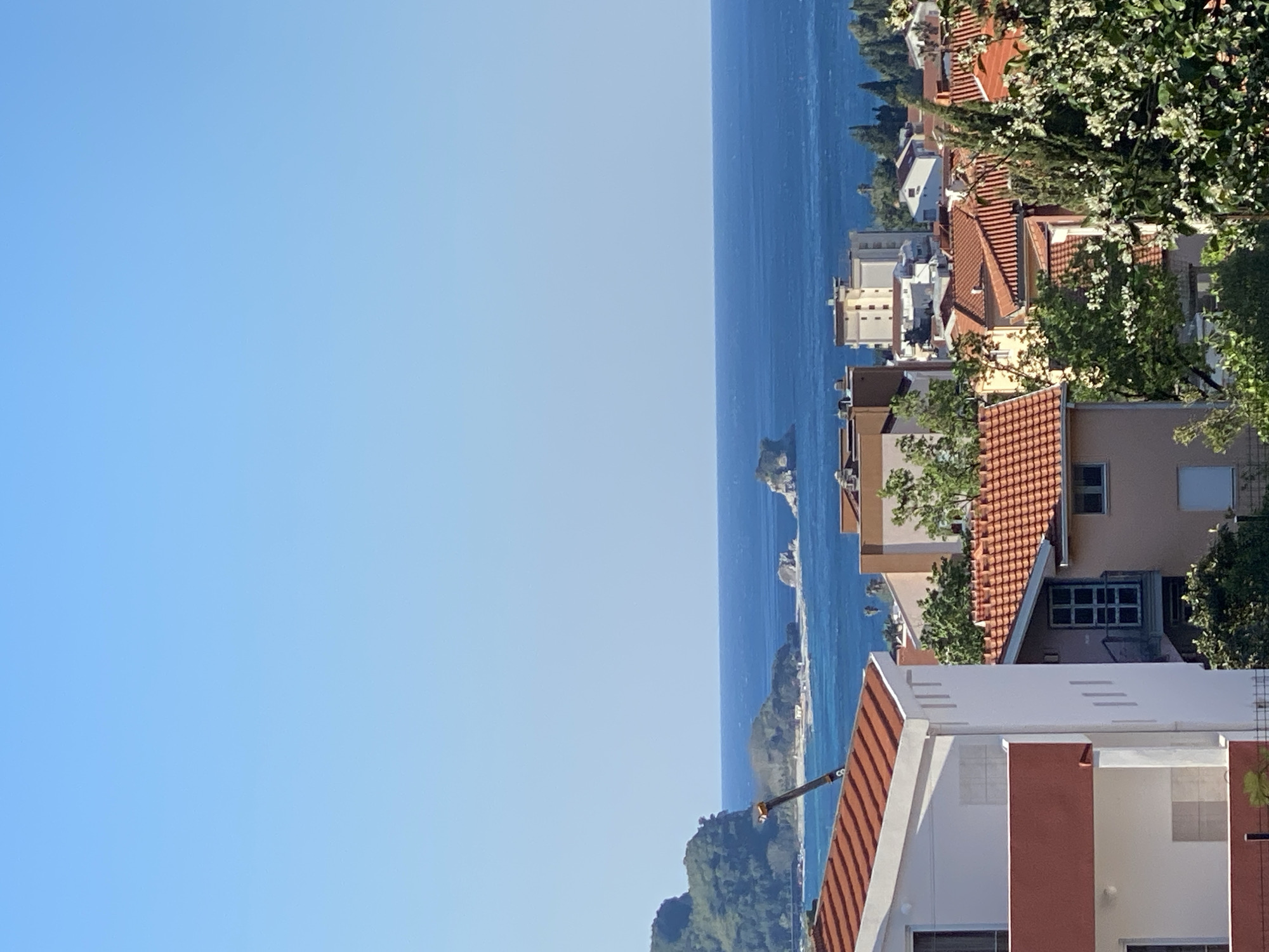 This media file presents a scenic, panoramic view of a coastal town and the sea, consistent with the provided context of Budva, Montenegro. The image appears to be taken from an elevated perspective, looking down towards the coastline and the expansive blue water.
The prominent subjects are the Adriatic Sea, the coastal architecture, and a clear blue sky. The sea is a deep, vibrant blue, stretching towards the horizon and featuring several small, rocky islets with sparse vegetation. The coastline is densely built, characterized by numerous buildings with distinctive red or orange terracotta tiled roofs. These structures vary in size and height, often featuring light-colored facades (white, beige, or pale yellow) and some with darker accents. Green trees and other foliage are visible throughout the townscape, interspersed among the buildings and on distant hillsides.
There are no people or specific activities depicted in the image; it is a static landscape photograph.
Notable details include the bright, clear blue sky, indicating excellent weather conditions. The strong, natural sunlight suggests it is daytime, likely morning or early afternoon. The architectural style, with its red-tiled roofs and coastal setting, strongly cues a Mediterranean location. The presence of rocky islets in the clear blue sea further supports the identification as the Adriatic coast. The overall impression is one of a beautiful, tranquil, and sunny coastal environment. There is no visible text in the image.