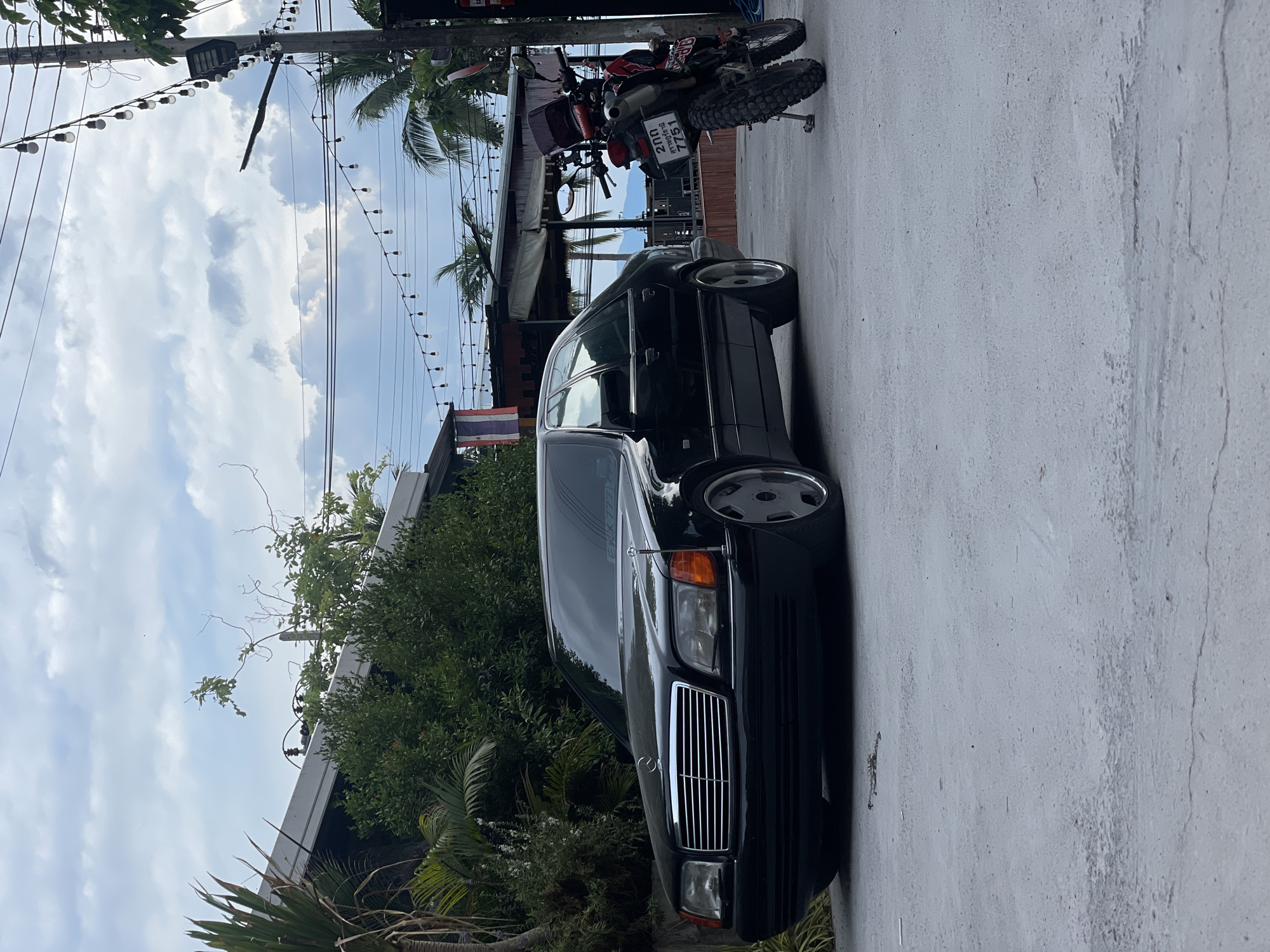 The image shows a black Mercedes-Benz parked on a concrete road. A motorcycle is parked further down the road, near a building with a partially visible awning. A Thai flag is visible in the background, near the building. Overhead, there are utility poles with wires and strings of lights. The sky is cloudy. Based on the license plate on the motorcycle and the presence of the Thai flag, the location is likely Baan Bang Rak, Thailand.
