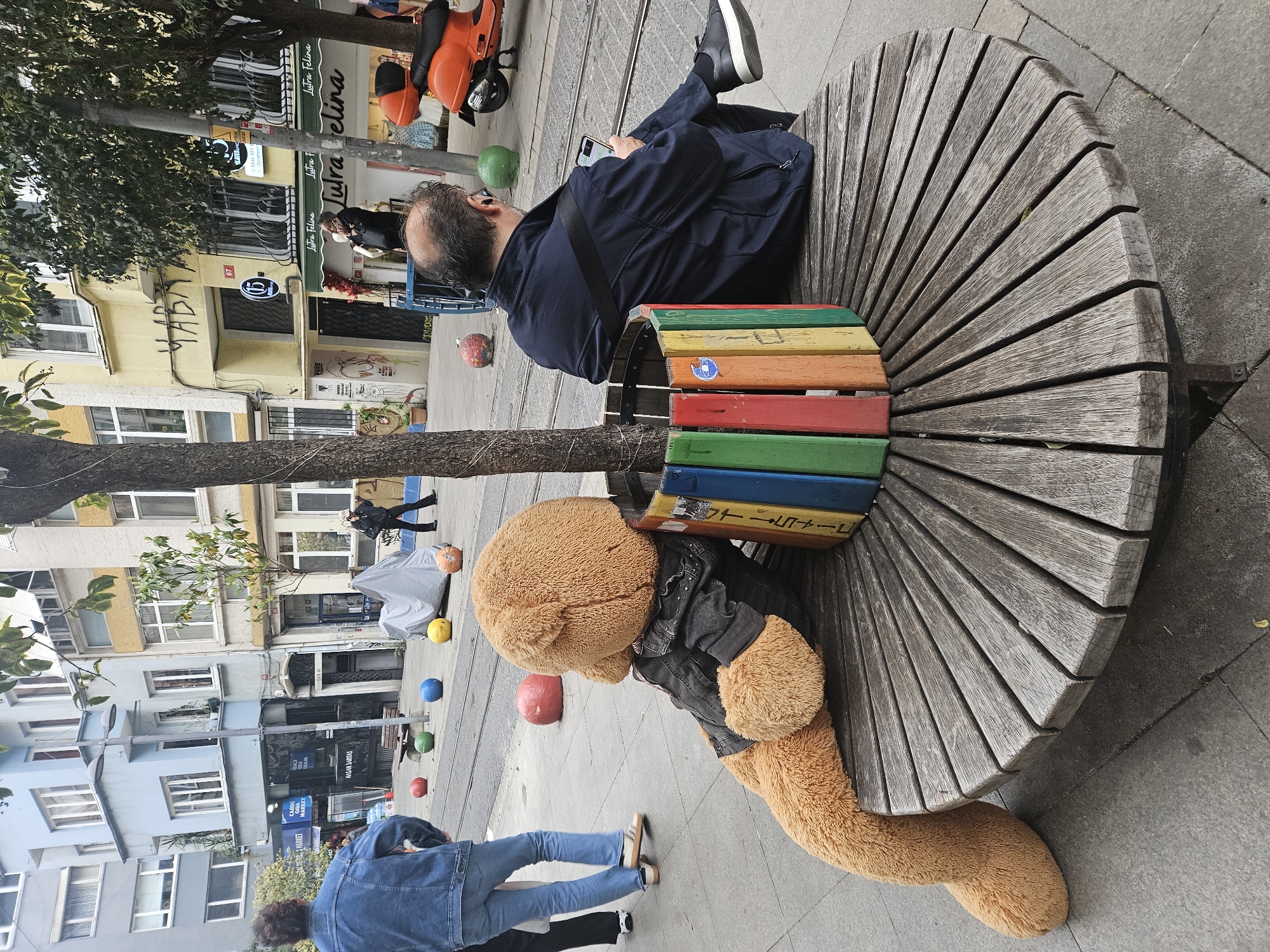 On a street in Kadıköy, Turkey, a large teddy bear in a black jacket rests against a round wooden bench wrapped around a tree, its legs extending onto the sidewalk. A man in a navy jacket sits on the bench, using his mobile phone. Buildings with businesses like "Lutra Felina" and "Cadde Market" line the street. Colored orbs dot the sidewalk. Pedestrians walk in the background. An orange scooter is parked nearby.