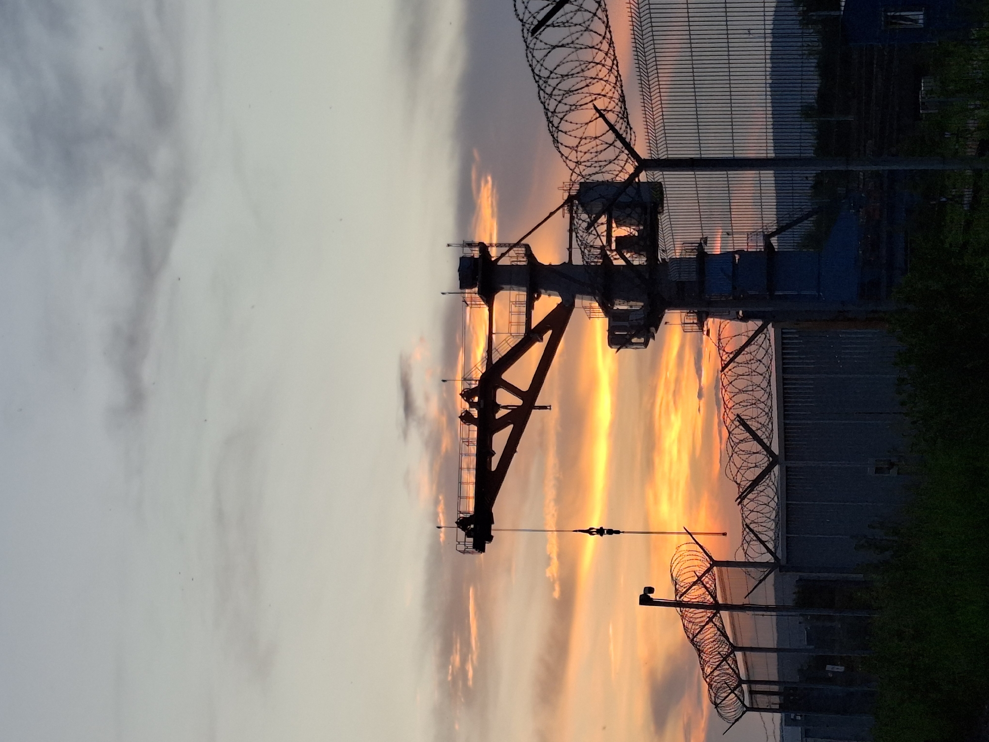 The image depicts a silhouetted industrial crane against a vibrant sunset sky over Chaikovsky, Russia. The sky is a gradient of orange, yellow, and grey clouds, with the sun setting behind a distant mountain range. In the foreground, a tall, dark industrial crane with a hook is positioned to the left of a cylindrical tower structure. A metal fence with barbed wire runs across the middle ground, separating the viewer from the industrial complex. Green foliage is visible at the bottom of the frame. The overall scene conveys an industrial atmosphere at dusk.
