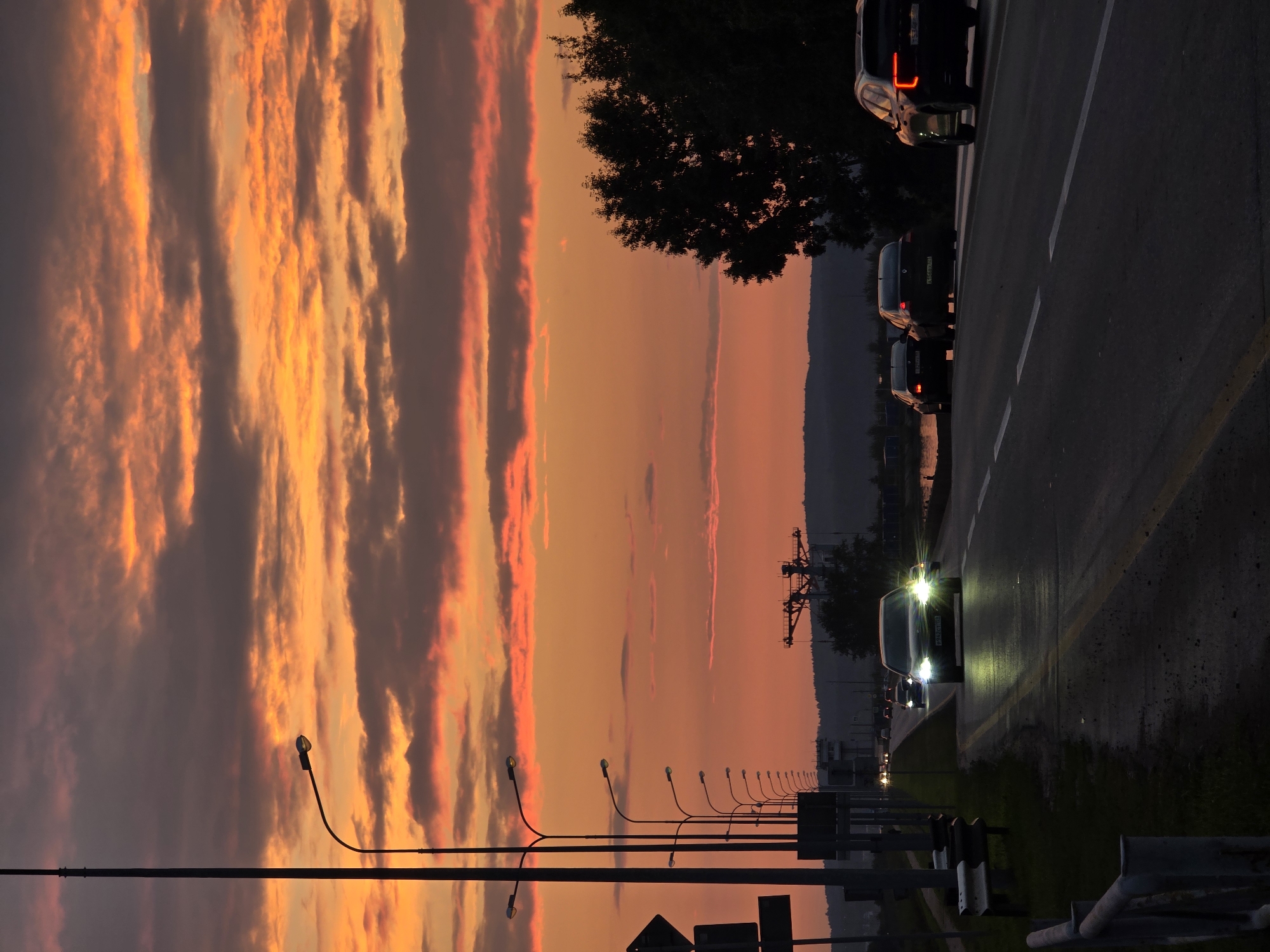 The image captures a twilight scene on a road in Chaikovsky, Russia, with a dramatic sunset painting the sky with vibrant orange and red hues. A line of streetlights stands silhouetted against the colorful sky on the left side of the road, casting a faint glow. Several cars are driving on the asphalt road, their headlights illuminating the path ahead. Some of these cars are in motion, suggesting traffic. In the background, trees and distant hills are visible, with a body of water appearing to the right. A large industrial structure, possibly a crane or part of a facility, is silhouetted against the horizon on the left. The road itself has clear lane markings, indicating it is a well-trafficked route. The overall atmosphere is serene yet dynamic, with the natural beauty of the sunset contrasting with the man-made elements of the road and vehicles.