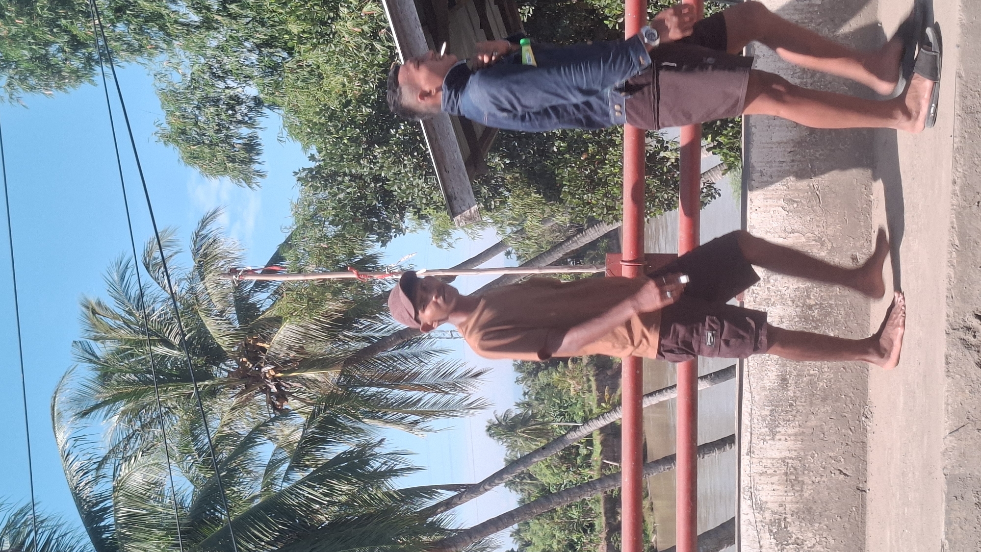 In the city of Kulukuta, Indonesia, two young men walk along a bridge. The man on the left, wearing a brown t-shirt and brown shorts, walks barefoot and appears to be looking towards the camera. He wears a brown baseball cap. The man on the right, in a blue denim jacket and dark shorts, walks with his head down and is smoking a cigarette. He wears a watch on his left wrist. The bridge has a red railing, and beyond it, a river is visible, lined with lush green trees and coconut palms. The sky is a clear blue. The walkway is made of concrete, and the scene appears to be in a rural or semi-rural area, characteristic of Kulukuta.