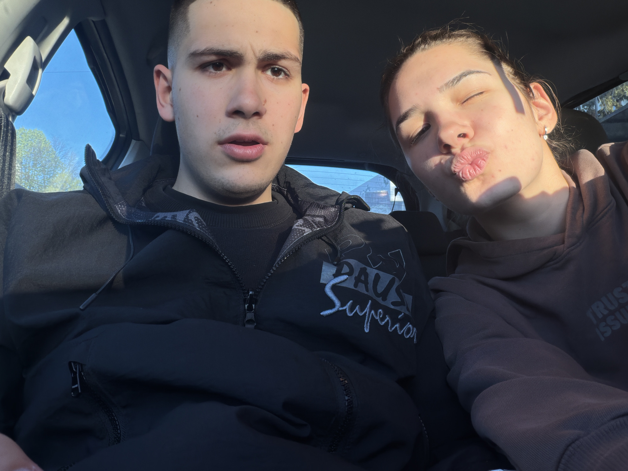 Two people are in the front seats of a car. A young man with short, dark hair is on the left, wearing a black jacket over a black sweater. The jacket has the logo "PAUL Superior" visible on it. He looks directly at the camera with a neutral expression. To his right, a young woman with her hair pulled back is making a kissing face, with one eye closed. She is wearing a brown hoodie. Sunlight streams in from the windows of the car. The background outside the windows is blurred, showing some green foliage and buildings. It is not possible to determine the precise location in Sofia, Bulgaria from the image.
