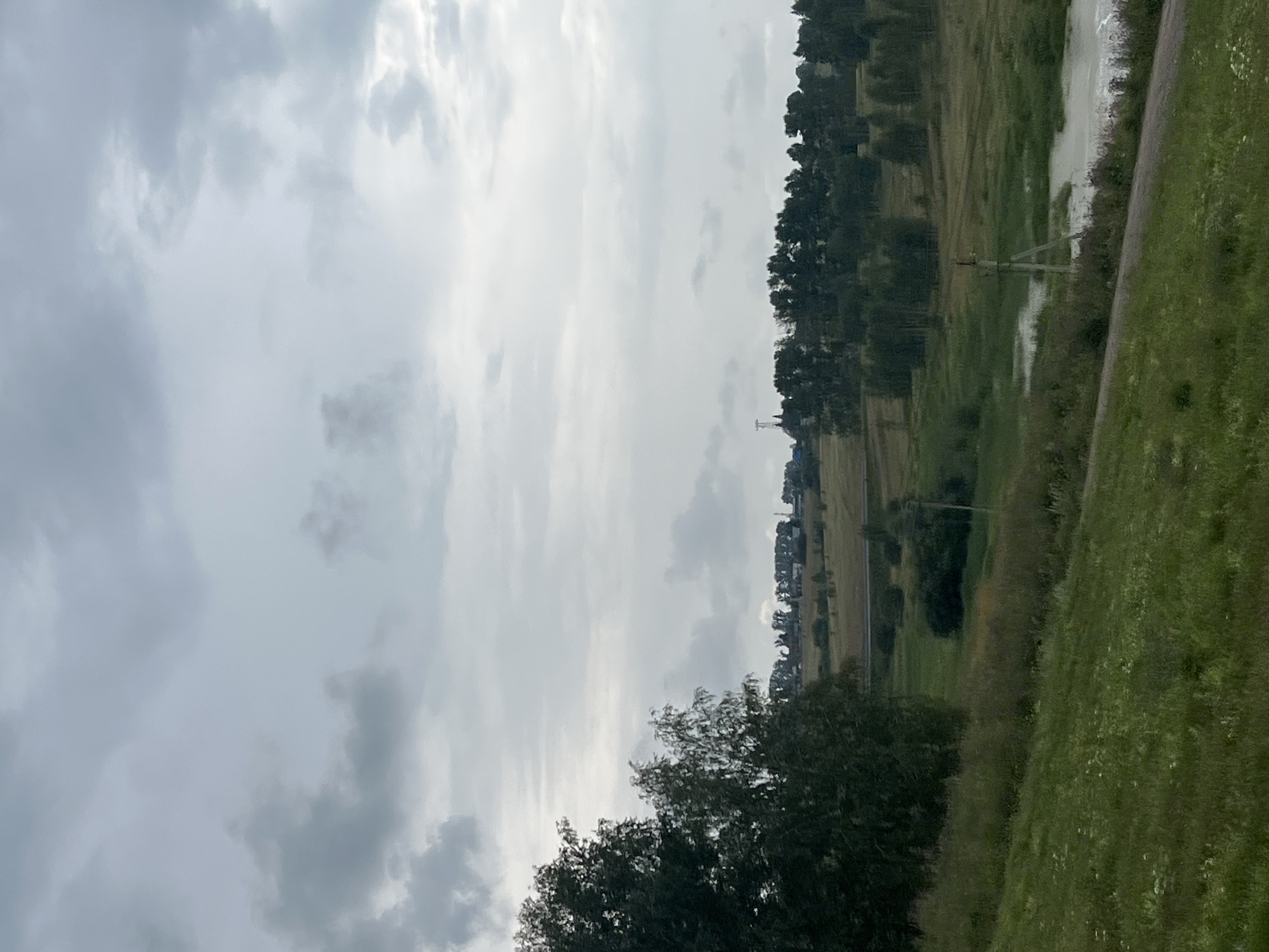 This image depicts a rural landscape under a cloudy sky. In the foreground, a grassy hill slopes downwards. Beyond the hill, a body of water, possibly a pond or slow-moving stream, is visible. A single utility pole with wires stands near the water. The middle ground is characterized by rolling fields and clusters of trees. In the distance, a line of trees forms a horizon, with hints of structures and a tall, slender tower visible above them. The overall environment suggests a natural, undeveloped area, consistent with the outskirts of Sokur, Russia. There are no people or animals visible in the image.