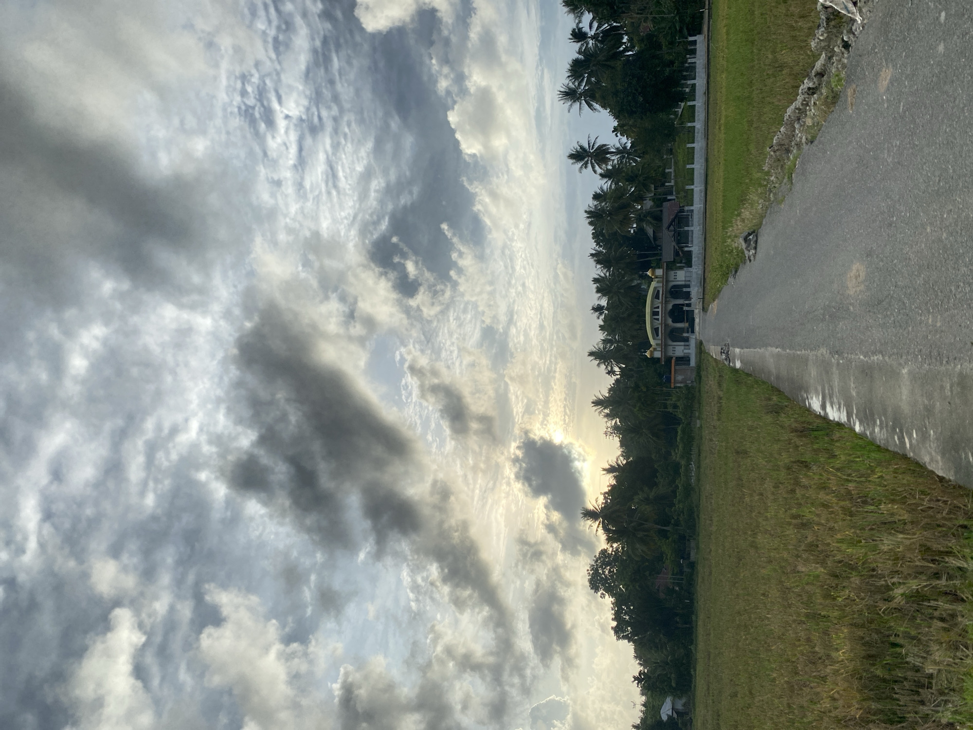 The image shows a landscape scene, potentially in Uteunreutoh, Indonesia. The sky is filled with clouds of varying shades of gray and white, with hints of sunlight filtering through. A line of trees, possibly palm trees, stands between the sky and the foreground. A building is visible among the trees. A road runs along the right side of the image, with green grass or vegetation on either side. The angle of the shot suggests the camera was tilted during capture.