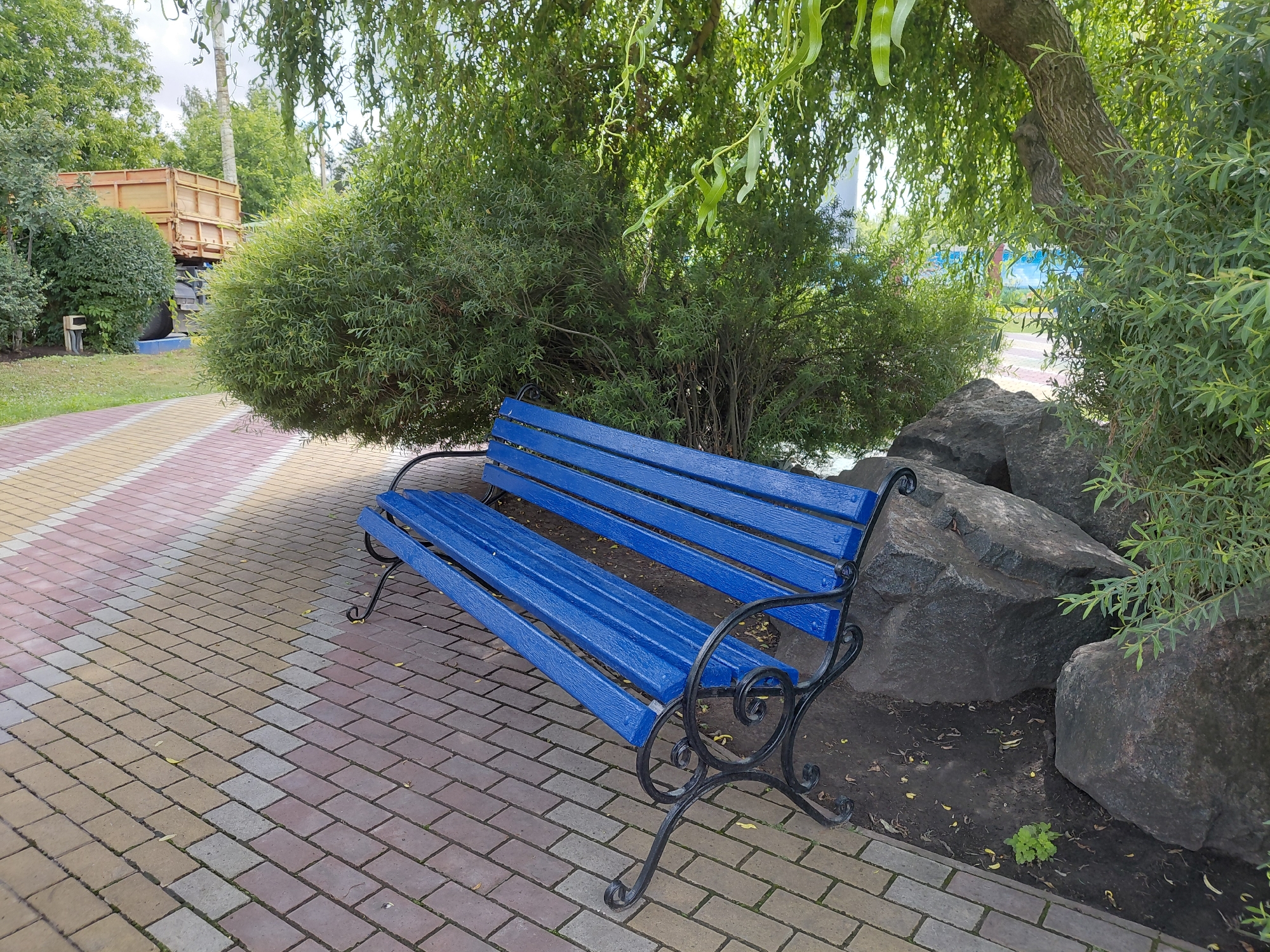 The image captures a vibrant blue park bench with ornate black metal legs, positioned on a decorative paved walkway. The walkway is composed of rectangular bricks in a repeating pattern of pink, beige, and gray. To the right of the bench, large, dark gray rocks are nestled amongst sparse greenery and fallen leaves. Behind the bench, a dense green shrub and the trunk of a mature tree with textured bark are visible, its branches draped with cascading green foliage. In the background, beyond the shrubbery, a large dump truck with a wooden bed is partially visible. The setting appears to be a park or a public outdoor space. The overall scene is tranquil and suggests a warm, possibly summer day. The location is identified as Kovalivka, Ukraine.