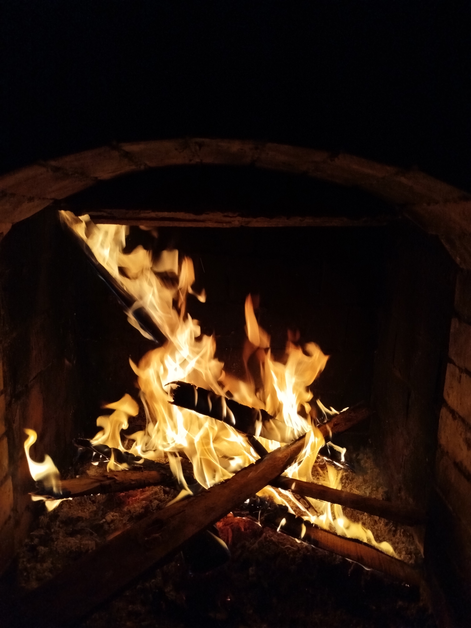 A nighttime view of a fire burning inside a brick fireplace. The fire is composed of burning wood and flames. The fireplace has an arched opening. The location is not identifiable as Lạc Dương, Vietnam.