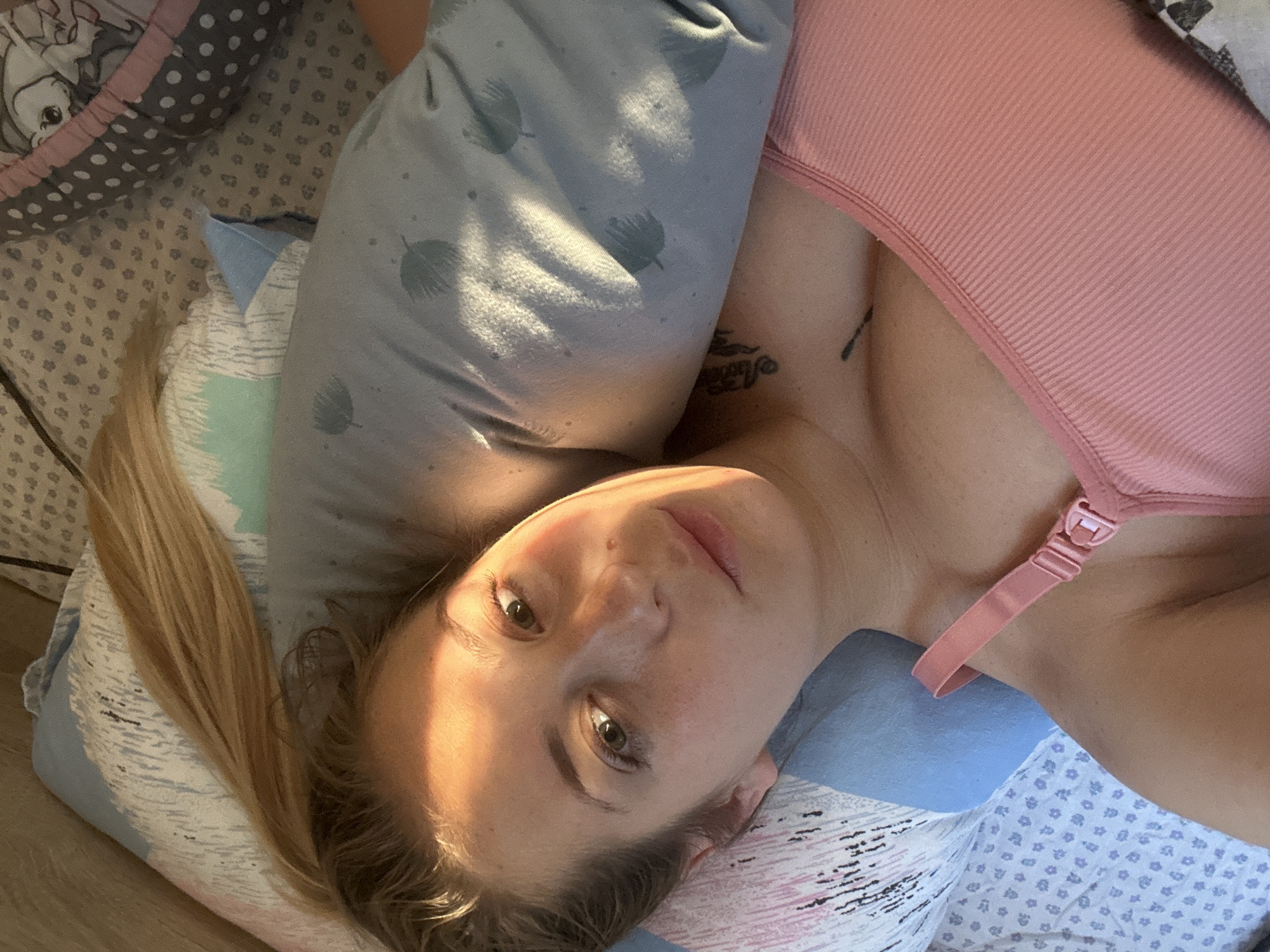 This media file shows a woman lying on her side in what appears to be a bed. She is wearing a pink ribbed tank top, possibly a nursing bra, with a clip visible on the strap. Her head is resting on a blue pillow with a feather-like pattern. A large grey pillow with a similar pattern is behind her. Her blonde hair is spread out.
The woman is looking upwards and to the side, with a relaxed or neutral expression. The lighting suggests it is daytime, with warm sunlight casting shadows on her face and the pillows. The background shows bedding with a pattern of small grey and white dots, and a glimpse of another patterned item, possibly a blanket or crib bumper.
The scene is domestic and intimate. There is a tattoo visible on her collarbone. No visible text is present in the image. The location context provided is Yavrovo, Bulgaria.