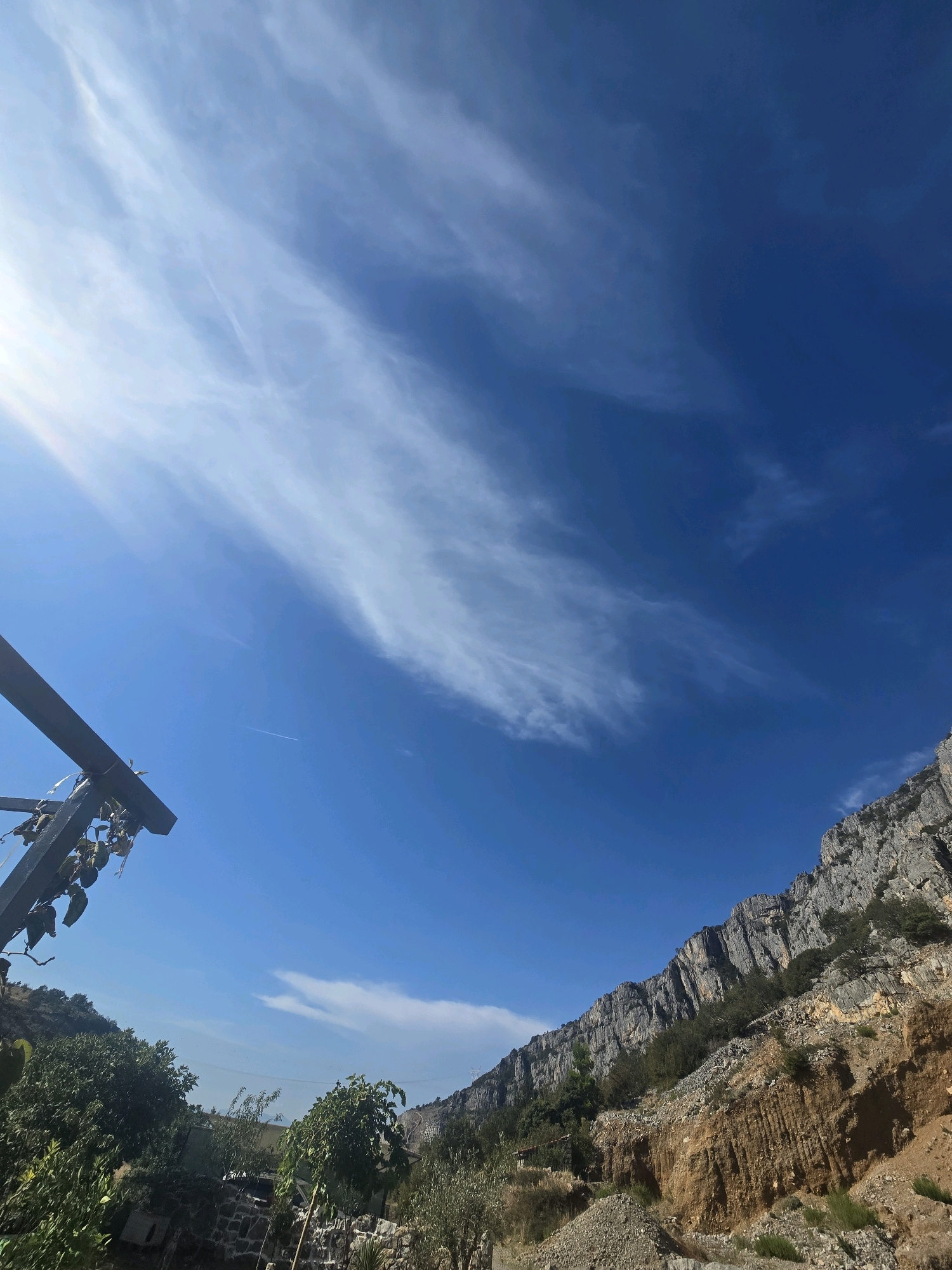 The image depicts a bright, sunny day with a clear blue sky dotted with wispy white clouds. A jagged, rocky mountain range dominates the right side of the frame, its steep slopes covered in sparse vegetation. In the foreground on the left, a dark, angular structure with some dangling objects extends into the frame, partially obscuring the sky. Below this structure, lush green trees and foliage are visible, suggesting a natural or semi-natural environment. The ground appears to be a mix of soil, gravel, and some stone-lined features. The overall impression is of a rugged, natural landscape bathed in strong sunlight, possibly on the outskirts of a city. While no specific landmarks or architectural styles definitively place this image in Podgorica, Montenegro, the mountainous terrain and vegetation are consistent with the region's geography. There are no people or specific actions captured in the image.