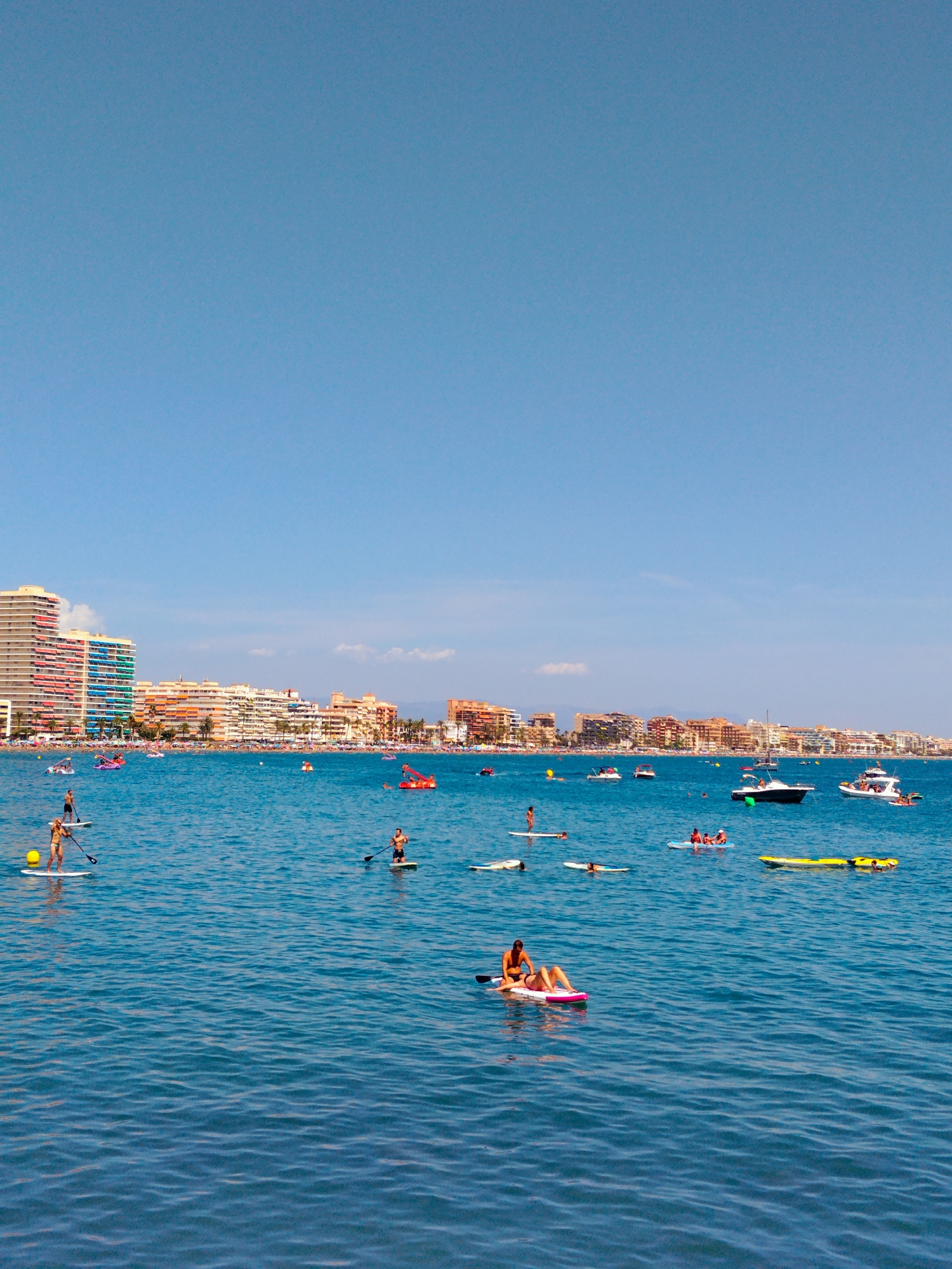 This is a wide shot of the sea with the cityscape of Peníscola, Spain, in the background. The water is a clear blue and calm, with small waves. Several people are engaged in various water activities. In the foreground, a man and a woman are on a paddleboard. To their left, a woman stands on a paddleboard. Further out in the water, another person is paddleboarding. A yellow inflatable raft is visible near the right side of the frame, with people on it.
In the mid-ground, several boats are present. One is a white motorboat with people on it. Another is a dark-colored motorboat. Further back, there are more boats and what appear to be inflatable water attractions.
The background features a dense cityscape with numerous buildings of varying heights and colors, primarily in shades of white, orange, and pink, suggesting a Mediterranean architectural style. A beach area with many people is visible between the water and the buildings. The sky is a clear, bright blue with a few wispy clouds. The overall scene depicts a vibrant and active summer day at a coastal resort.