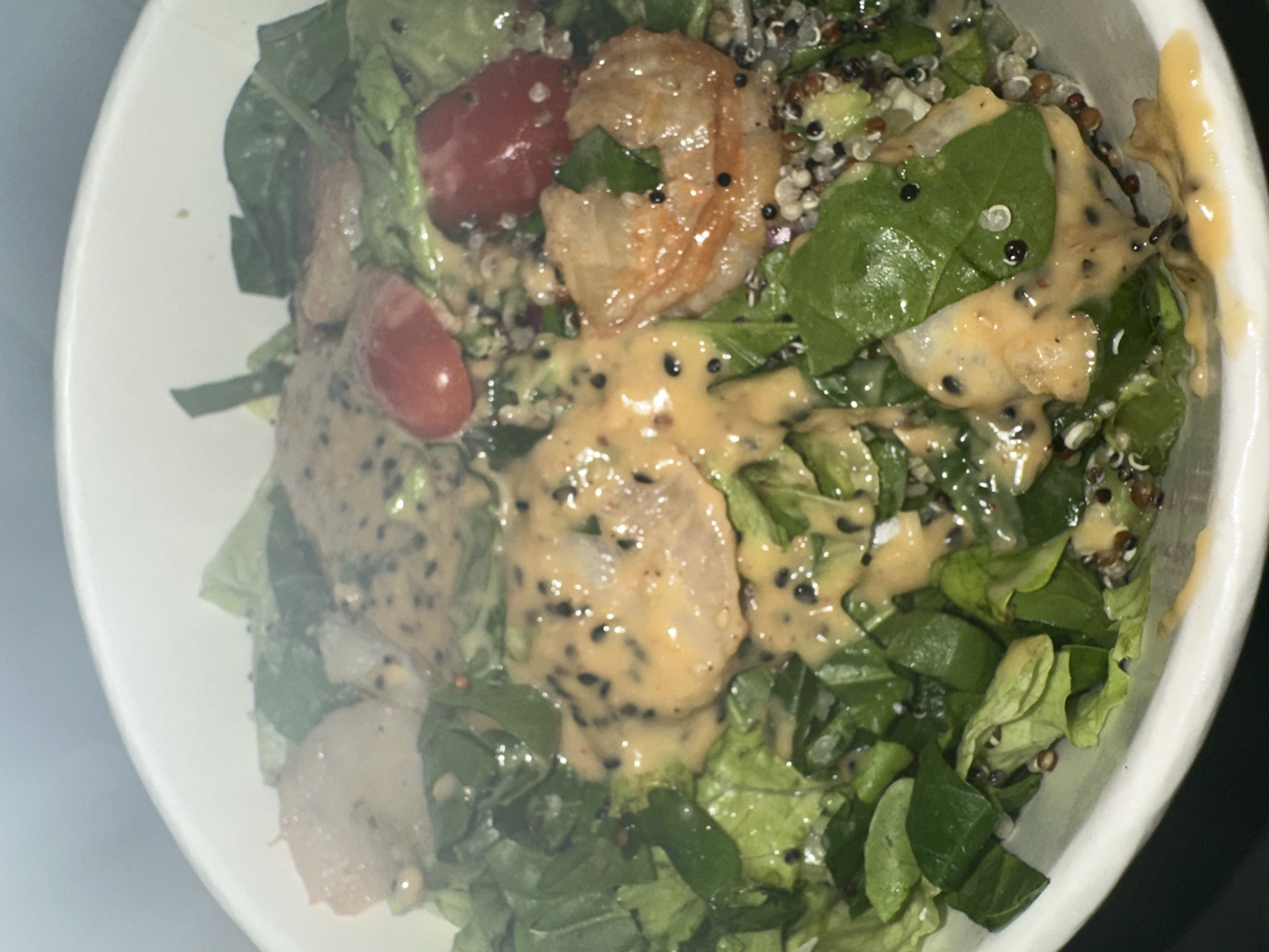 A close-up, high-angle shot shows a salad in a white bowl. The salad contains mixed greens, possibly including spinach and lettuce, interspersed with what appear to be cooked shrimp and halved cherry tomatoes. A beige-colored dressing, speckled with black seeds, is generously drizzled over the salad. The overall impression is a fresh salad with protein and a creamy dressing. There is no information to suggest this picture was taken in Sofia, Bulgaria.