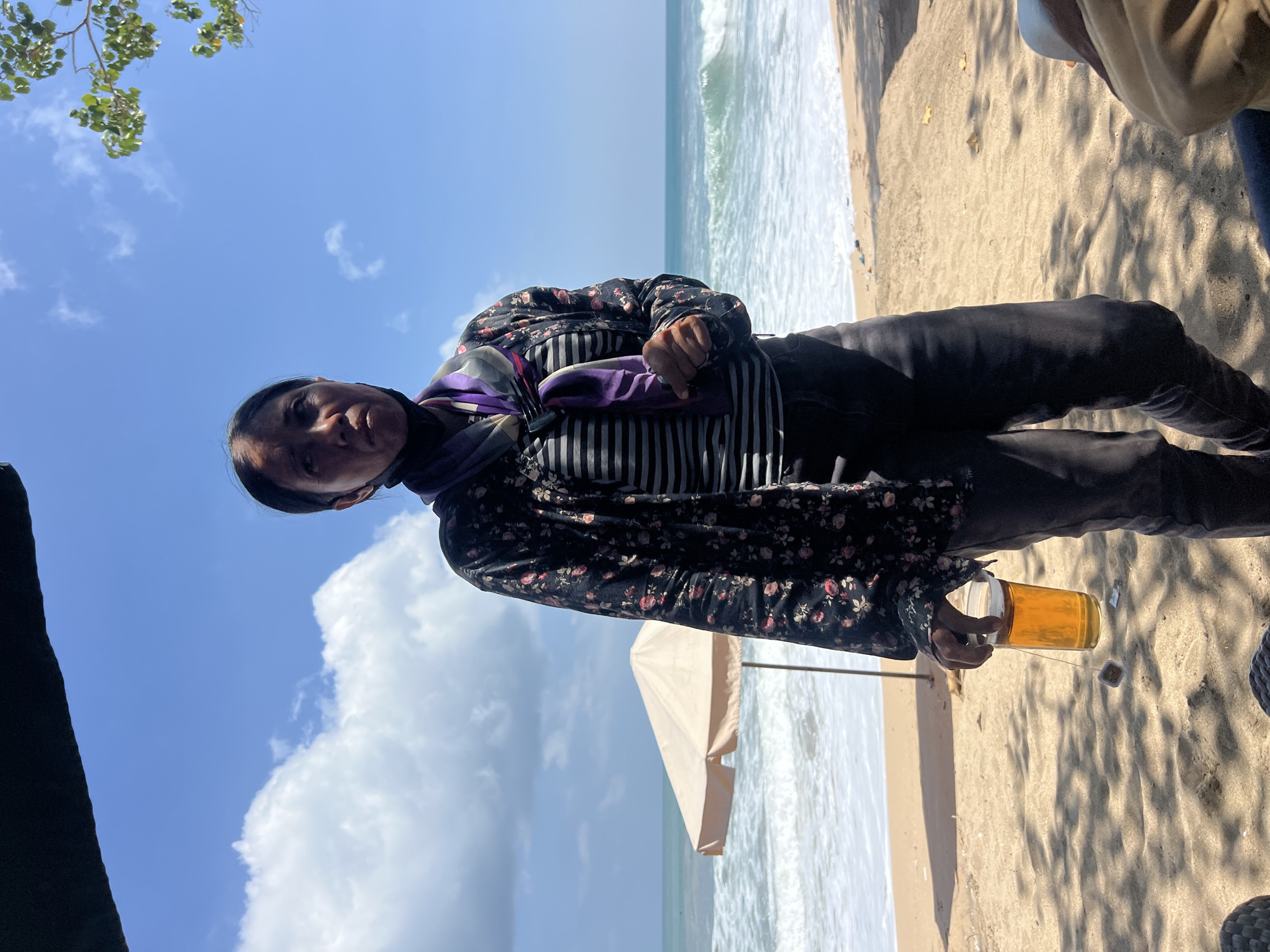 A person stands on a sandy beach in Kuta, Indonesia, holding a clear plastic cup filled with amber liquid and a tea bag string. The individual is wearing a black floral print jacket over a striped shirt and dark pants. A purple and white scarf is wrapped around their neck, and a black mask is worn below their nose. The background features the ocean with moderate waves, a cream-colored beach umbrella, and a clear blue sky with scattered white clouds. Trees with green leaves are visible in the upper right corner, casting shadows on the sand. The overall scene depicts a casual outdoor setting on a sunny day.