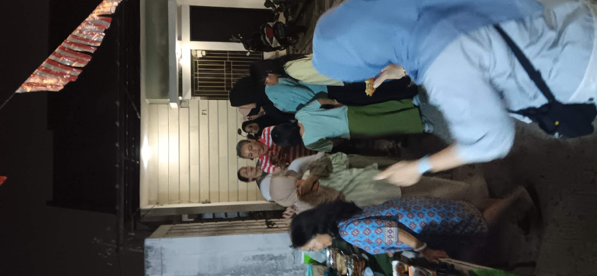 The image depicts an outdoor gathering at night, likely in a residential area of Depok, Indonesia, given the style of the house and the presence of a motorcycle. Several people, including women and a young girl, are gathered around a food stall or table laden with various dishes. The atmosphere appears casual and convivial, with some individuals engaged in conversation and others observing the food. Decorative banners, possibly for a celebration, are visible hanging from the roofline. The lighting suggests it is evening, with artificial lights illuminating the scene. The foreground is partially obscured by a person wearing a blue headscarf, who is holding a piece of food and gesturing with their other hand.