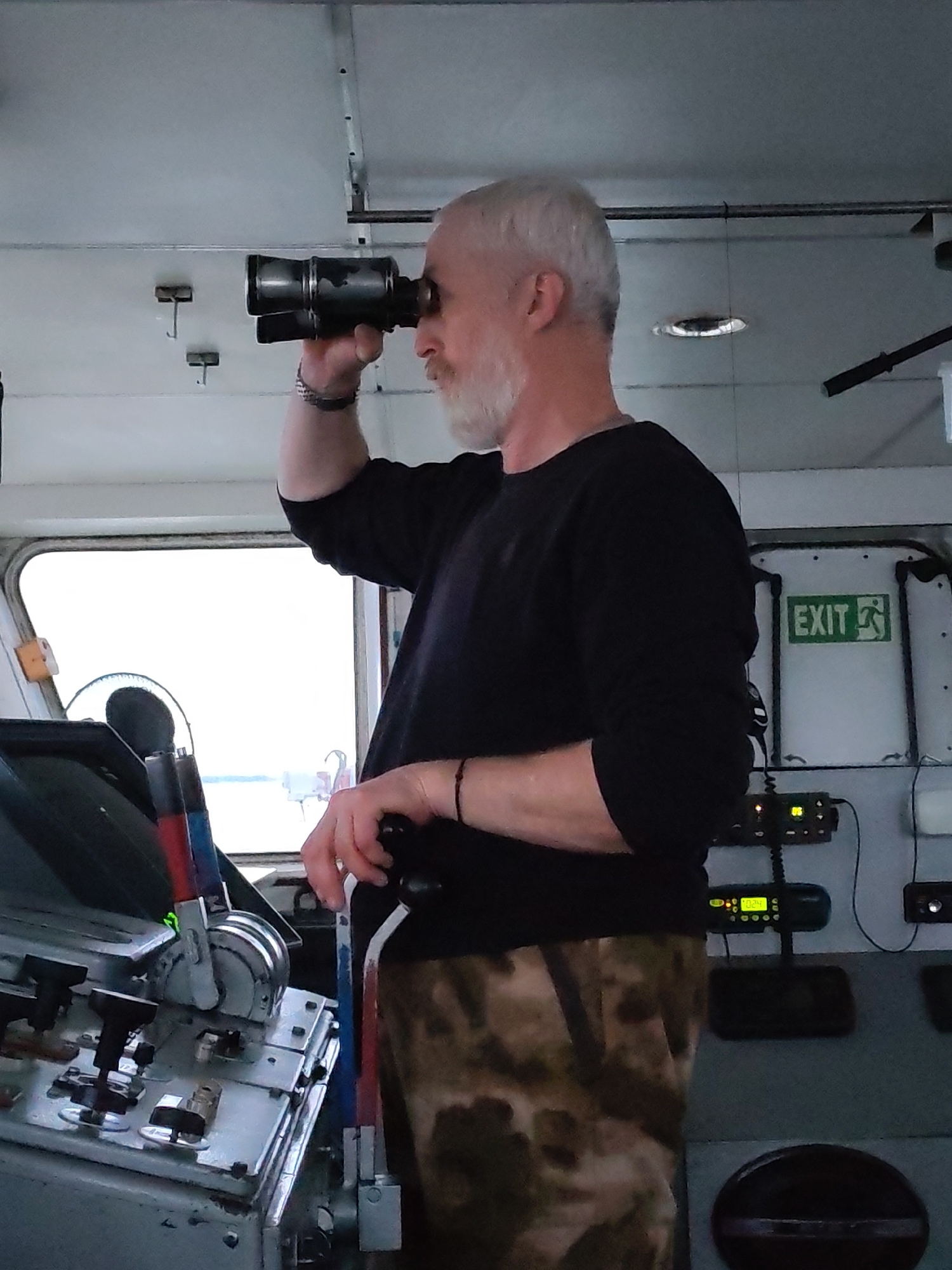 A man with white hair and a beard is standing on what appears to be the bridge of a ship. He is holding binoculars to his eyes and looking out of a large window. The bridge is cluttered with equipment, including a control panel with levers and buttons, and radio equipment. An exit sign is visible on the wall. The location in the city is unknown.