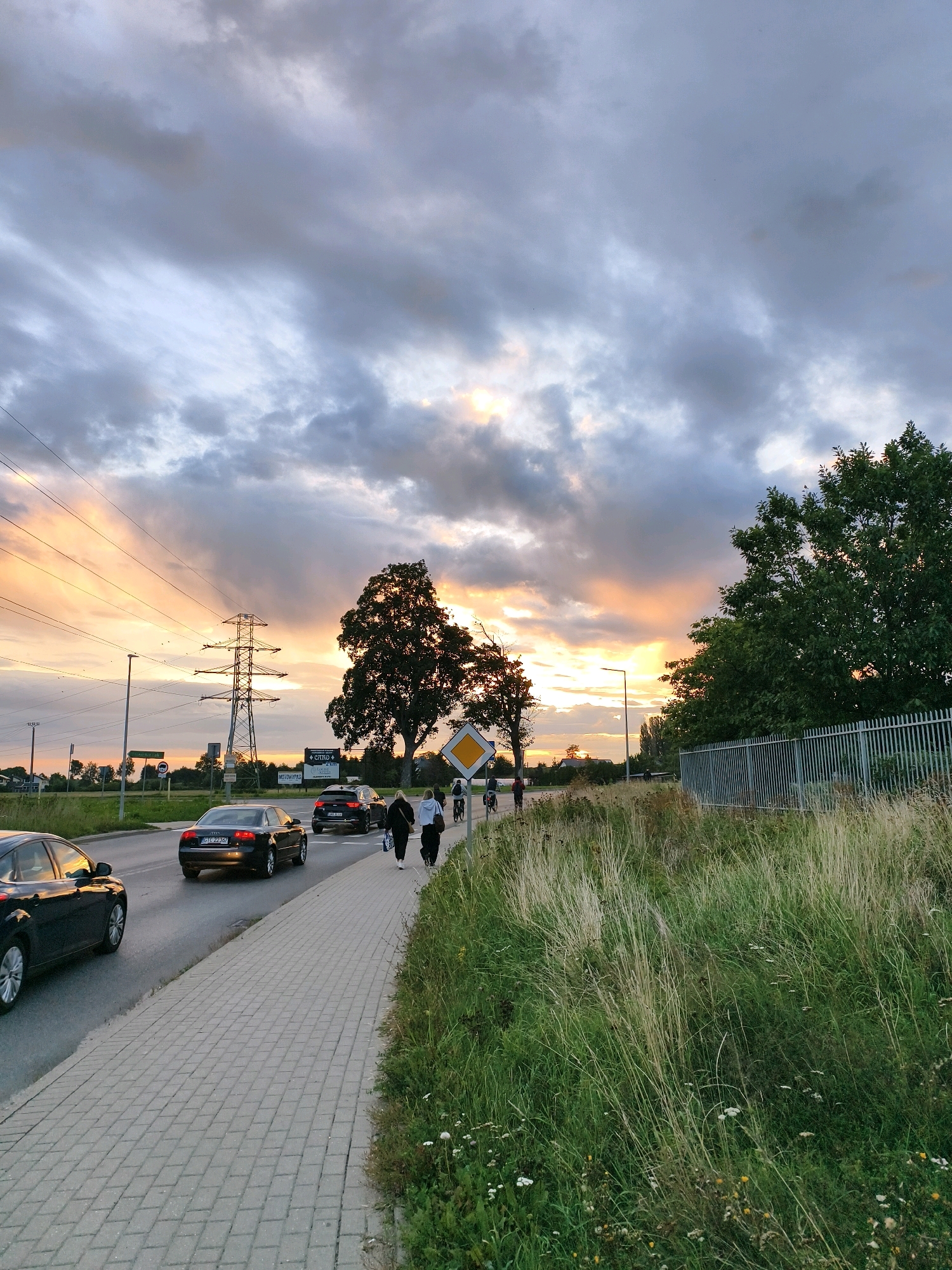 The image captures a street scene in Tczew, Poland, during sunset. A paved sidewalk runs along the right side of a road with vehicular traffic. Several cars are visible on the road, including a black Audi A4 with license plate GTC 22397 in the foreground. On the sidewalk, three individuals are walking away from the camera, two appearing to be women and one a cyclist. To the right of the sidewalk, tall grass and a metal fence line the area. Across the road, a large tree stands prominently, with electrical pylons and power lines in the background, suggesting an urban or suburban environment. The sky is filled with dramatic clouds, illuminated by the warm hues of the setting sun.