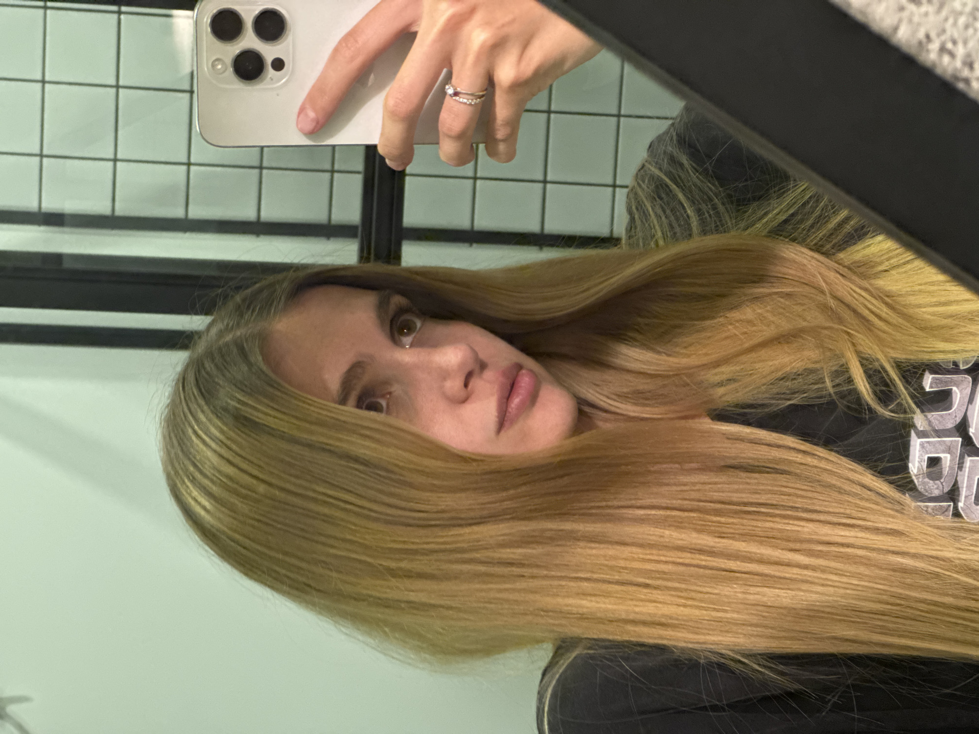 A young woman with blonde hair is taking a selfie in a mirror. She is holding a white smartphone with three rear cameras in her right hand, which is visible in the upper part of the frame. A ring is on one of her fingers. The background behind her includes a wall with square tiles and black horizontal bars. She is wearing a black garment. The scene appears to be in a modern-style bathroom or similar setting with light green walls and a tiled wall. It's impossible to determine the exact location in Tbilisi, Georgia, without additional information or context.