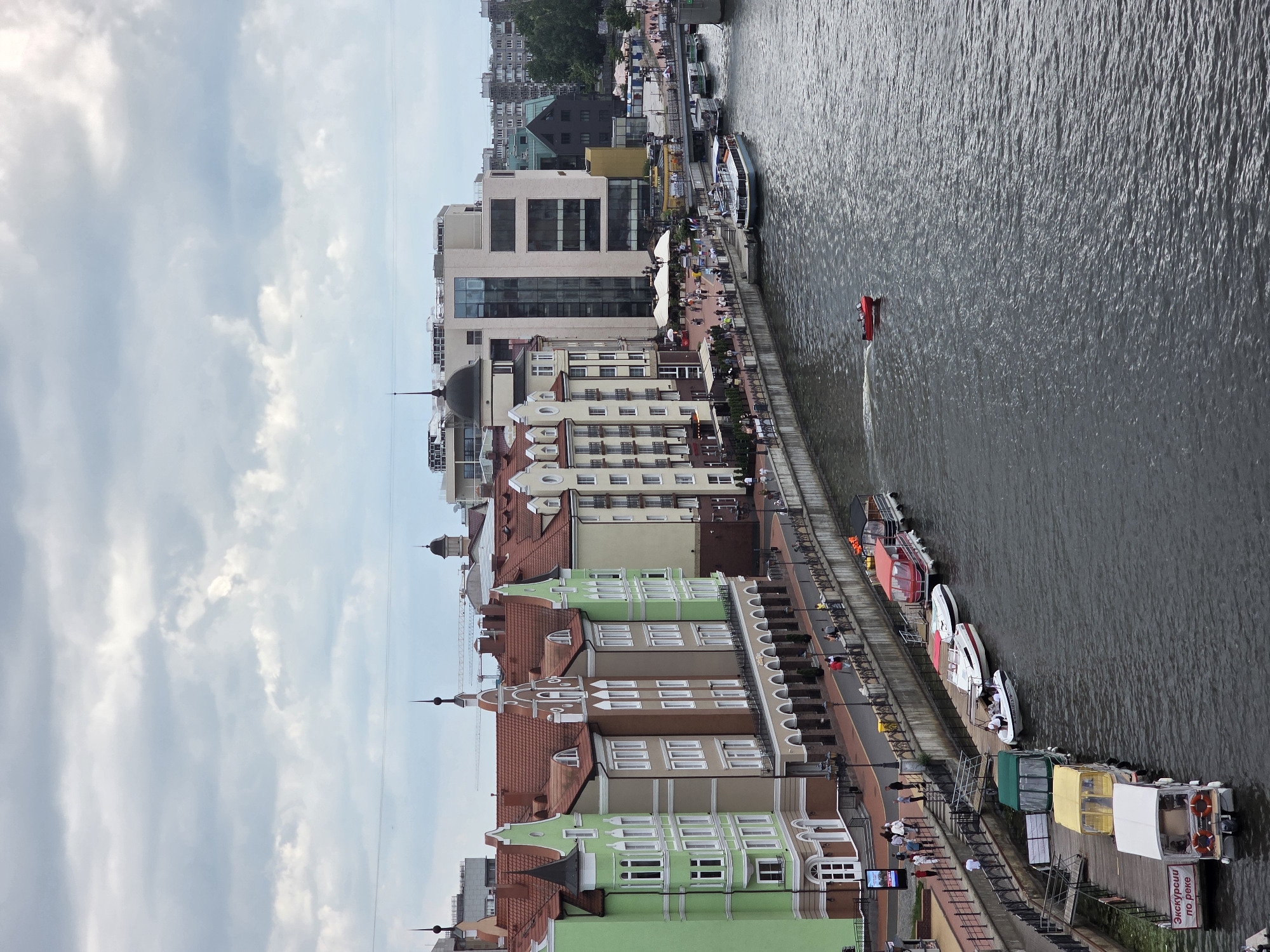 The image captures an elevated view of the Pregolya River embankment in Kaliningrad, Russia, specifically focusing on the area known as "Fish Village" (Рыбная деревня).
The foreground features the dark, rippled surface of the Pregolya River, flowing from the right to the left of the frame. A small red speedboat with at least two individuals onboard is visible mid-river, moving towards the right and generating a white wake.
Along the immediate left bank of the river, a stone retaining wall supports a paved promenade. A black metal railing with decorative lampposts borders the river, along which numerous pedestrians are walking. Behind the promenade, a row of multi-story buildings with a distinctive architectural style characteristic of "Fish Village" extends. These buildings feature prominent pitched, reddish-brown tile roofs, gabled facades, and multiple white-framed windows. From left to right, their primary exterior colors are light green, beige, reddish-brown, and pale beige/white. The ground floors of these structures incorporate arched walkways or colonnades.
Further along the left bank to the right, beyond the historically styled buildings, modern multi-story commercial and residential buildings are visible, including a prominent light-colored building with large dark-tinted windows. Outdoor seating areas with white market umbrellas are present on terraces along this section, with people occupying them.
Multiple boats are docked along the left bank's wooden piers and stone moorings. These include several white motorboats, one red-canopied boat, one green-canopied boat, and a small white passenger boat with a sign in Russian reading "ЭКСКУРСИИ по реке" (EXCURSIONS by river). Additional larger passenger vessels are visible further downriver, near a partially obscured bridge on the far right.
The opposite bank of the Pregolya River displays a mix of urban structures, including multi-story residential buildings and some industrial or commercial buildings, interspersed with green tree cover. Two construction cranes are discernible against the distant skyline on the far bank.
The sky is heavily overcast with dense, light-grey and white cloud formations, indicating a cloudy daytime environment.