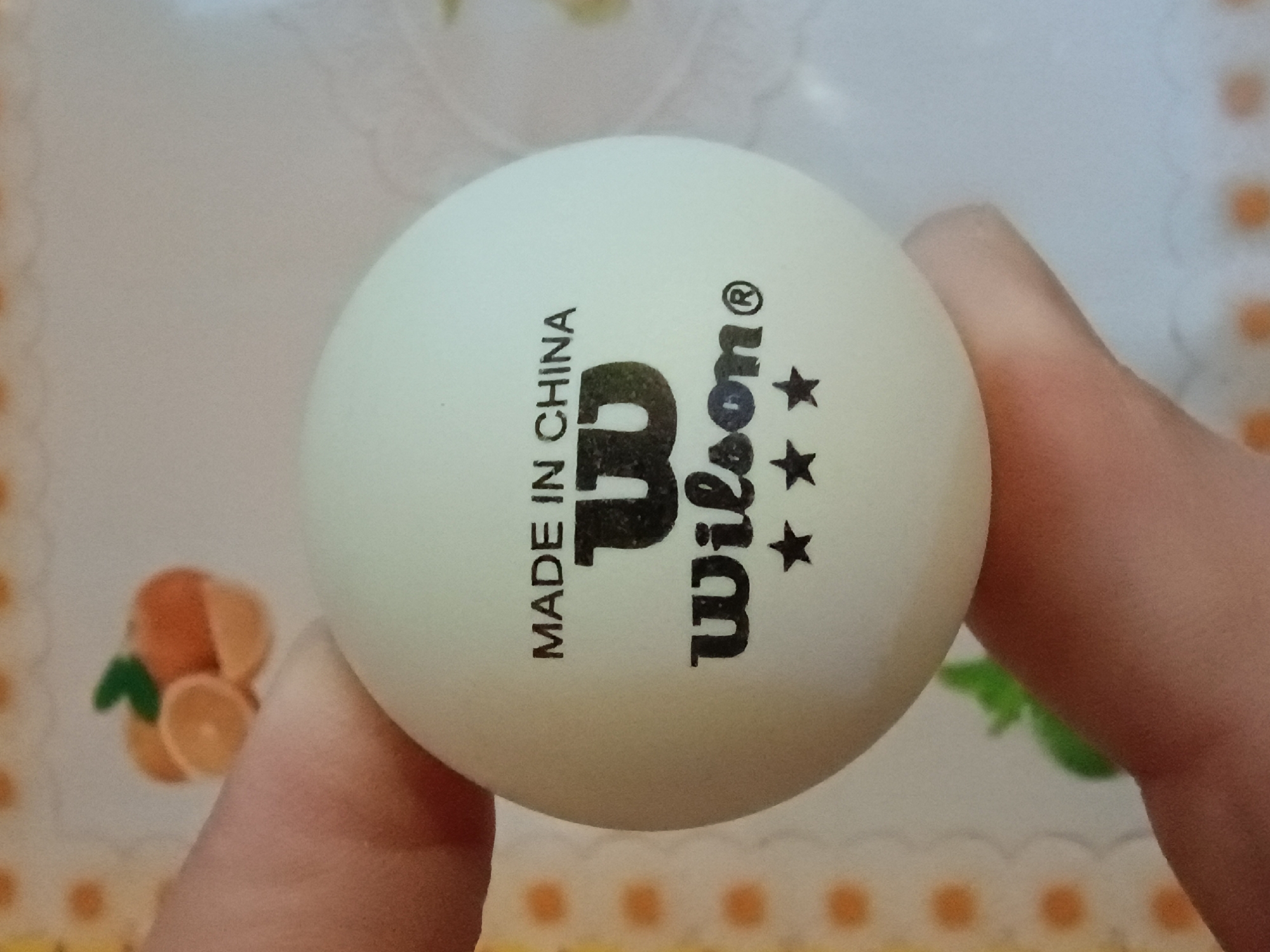 A white ping pong ball with "MADE IN CHINA" and the "Wilson" logo printed in black is held by two fingers. Three black stars are printed below the logo. The background is a light-colored surface with a decorative pattern of yellow circles and white scalloped edges. A blurred image of oranges and a green leaf is visible in the upper left corner.