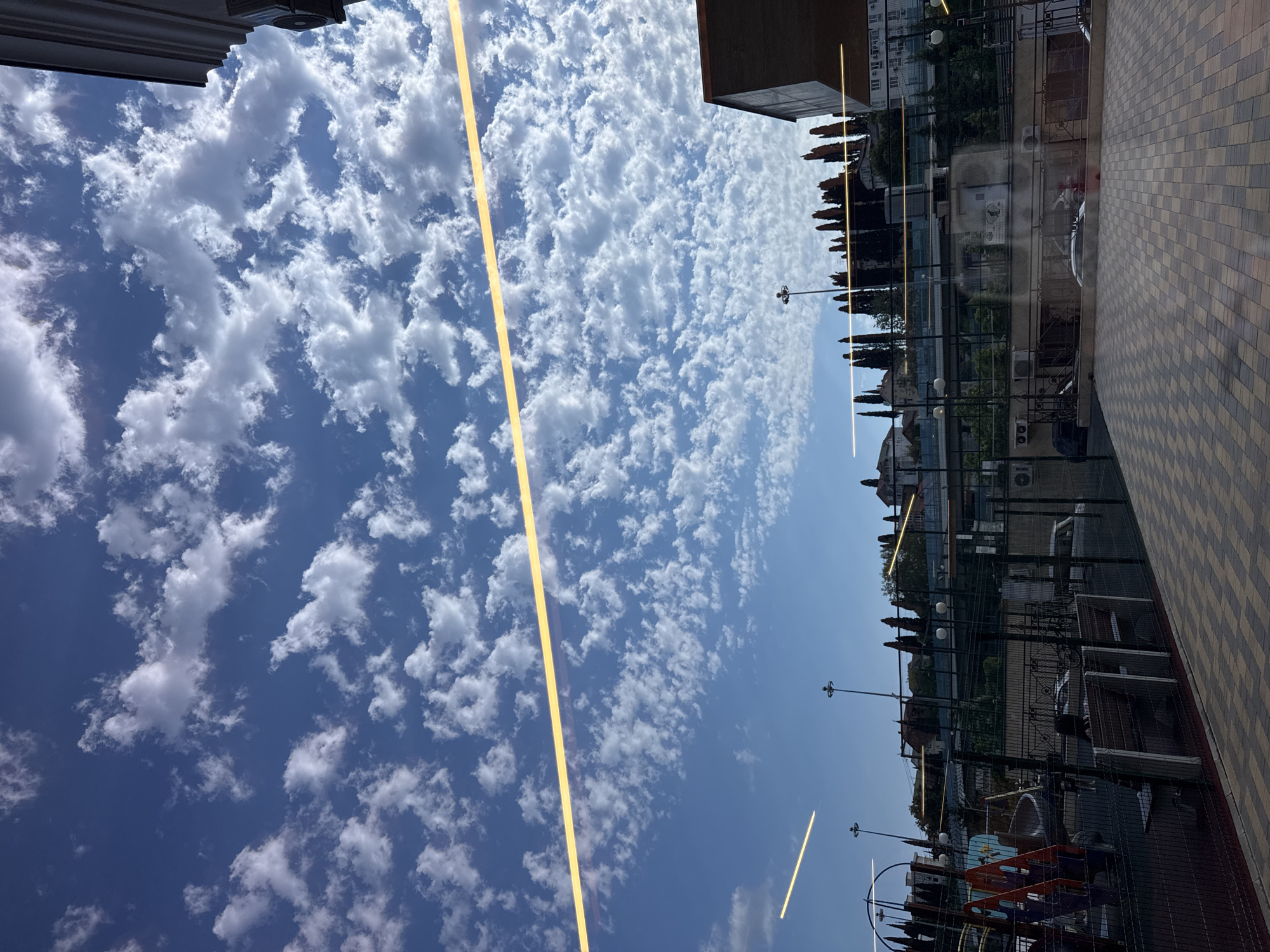 The image shows a view of a street scene, possibly a promenade or courtyard, with buildings and trees lining one side and a paved walkway on the other. The sky is a clear blue, dotted with fluffy white clouds, suggesting it is daytime and the weather is pleasant. A thin, bright yellow line, possibly a light fixture or reflection, runs diagonally across the image, separating the sky from the lower portion. In the background, several buildings, some with balconies and windows, are visible, along with tall, slender trees. The paved walkway features a checkerboard pattern of beige and brown tiles. There are streetlights and some outdoor seating or play equipment visible. The scene appears to be set in an urban or resort area, given the presence of buildings, paved areas, and decorative trees. The rotation of the image suggests it may have been taken on a phone and not oriented correctly. No people are clearly visible, and the overall mood is serene and bright.
