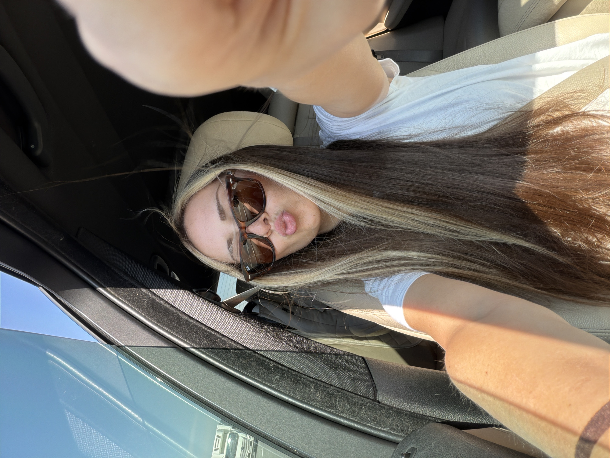 The image shows a selfie taken by a person inside a car. The person is wearing sunglasses and has long brown and blonde hair. They are wearing a white shirt. The interior of the car is visible, including the seat and window. The specific location in Moscow, Russia, cannot be determined from the image.