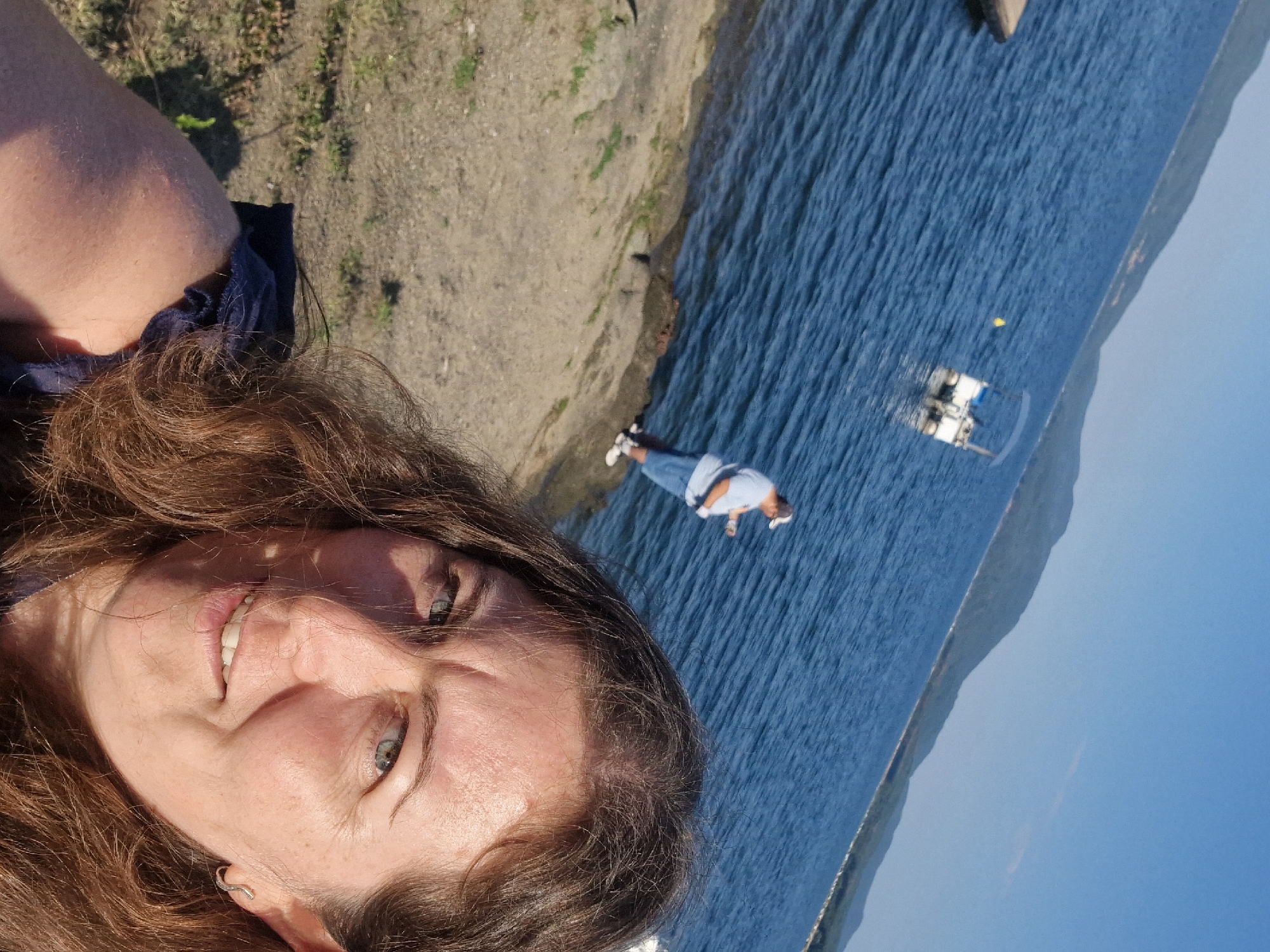 In the city of Dolni Pasarel, Bulgaria, a medium shot captures a woman in the foreground looking towards the camera, with a lake and distant hills in the background. The woman has long, wavy brown hair and is wearing a dark blue top. Her face is lit by sunlight, highlighting her blue eyes and a slight smile. She has on a silver hoop earring. In the mid-ground, a man is standing by the water's edge, appearing to be fishing. He is wearing a light blue t-shirt, blue shorts, a white baseball cap, and white sneakers. A small white boat with a canopy is moored in the water behind him. The water is a deep blue, and the shore is rocky and sandy, with sparse vegetation. The sky is clear and blue, with distant, tree-covered hills forming the horizon. The scene conveys a sense of a peaceful day by the lake.