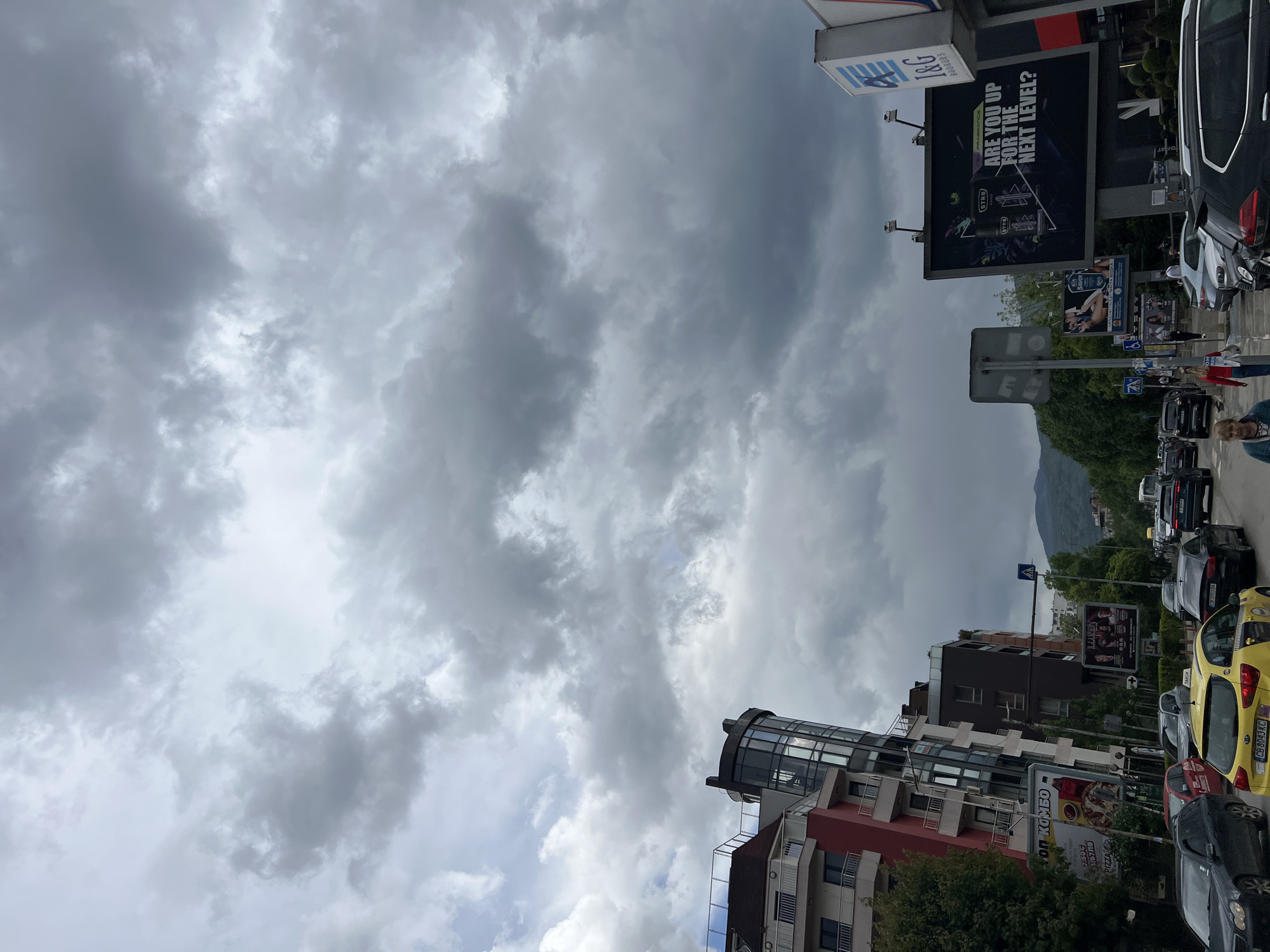 The image depicts an urban scene with a cloudy sky dominating the upper half of the frame. Two large billboards are visible on either side of the sky. The left one has "T&G Brokers" on it and the other has "ARE YOU UP FOR THE NEXT LEVEL?".
The lower half of the image shows a street scene with buildings, cars, and signs. A multi-story building with a circular glass structure on top is located on the right side. Various advertisements and signs are attached to the buildings, including one promoting pizza. Cars are parked and lined up along the street, a yellow taxi being one of the most prominent.
The location is likely in Sofia, Bulgaria, indicated by the car registration plate "CB 6043KC" on the yellow taxi. A mountain is visible in the background.