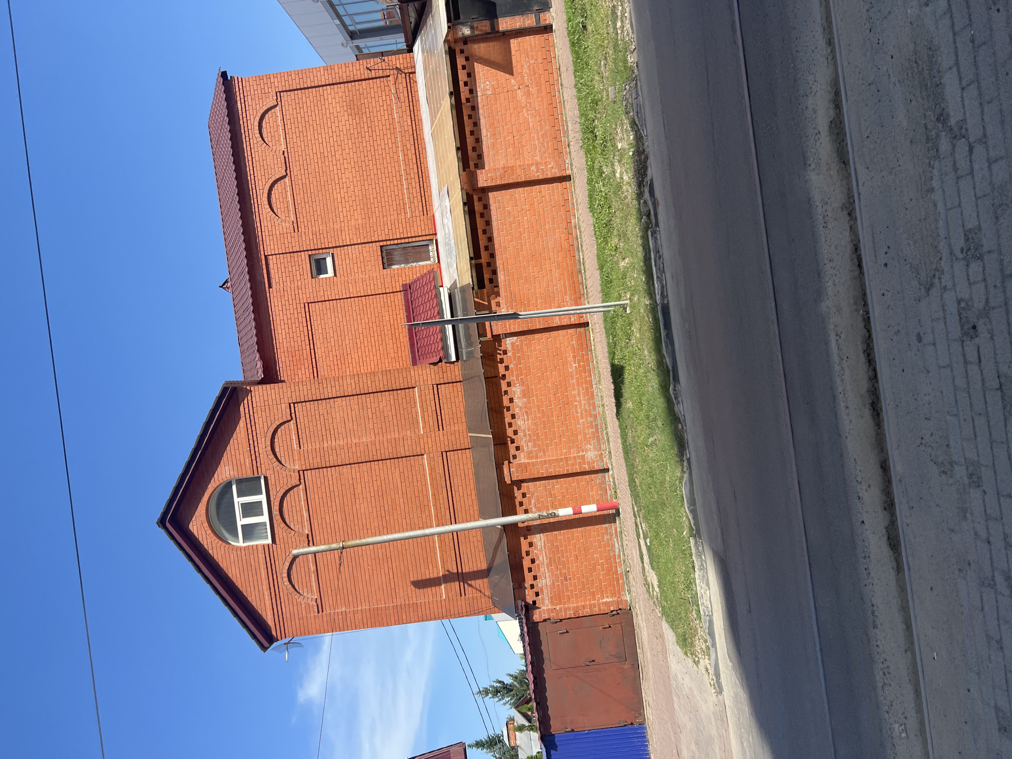 The image shows a brick building and a brick fence along a street. The building is red brick with a dark red roof and a few small windows. There are arched decorative elements on the side of the building. A metal pole with red and white markings stands in front of the building. To the left of the building is a rusty metal garage door and a blue metal fence. The street in the foreground is asphalt with a concrete sidewalk and grass. The sky is clear and blue. The image was taken in Ulyanovsk, Russia.