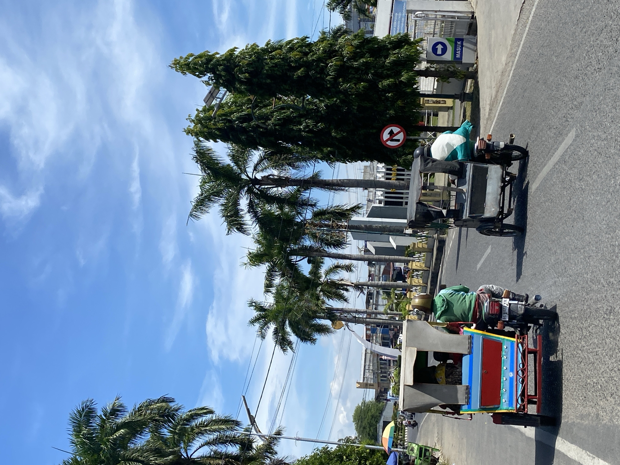 In Lhokseumawe, Indonesia, under a bright blue sky with scattered clouds, two three-wheeled vehicles, commonly known as becaks, traverse a paved road. The becak in the foreground is vibrantly colored in red, blue, and yellow, with a beige canopy. It is ridden by a person wearing a green hooded jacket and gray pants, their face obscured by a helmet. The becak behind it is darker in color with a gray canopy and is driven by a person wearing a turquoise garment with a white pattern, also wearing a helmet. Palm trees and tall, slender trees line the road, with buildings visible in the background. A traffic sign with a red circle and arrow indicates no right turn, and a blue sign with an arrow and the word "MASUK" (which means "ENTER" in Indonesian) is posted near a gate. The scene captures a slice of everyday life and transportation in a city in Indonesia.