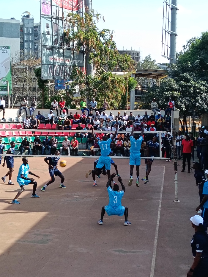 An outdoor volleyball match is in progress on a reddish-brown hard court at a sports complex in Nairobi, Kenya. Approximately seven male players are visible on the court. Three players in light blue jerseys (numbered 1, 3, and 11) are actively engaged in play. Player 11 and an obscured teammate in light blue are airborne near the net, poised for a block or spike, while player 3 is in a low, ready stance. A player in a dark blue jersey is in an airborne, bent-over position near the net, possibly reacting to the ball. A yellow and blue volleyball is suspended in mid-air above the net.
Tiered spectator stands with red and green plastic seating are visible in the background, occupied by numerous onlookers, predominantly male. Above the stands, some individuals are seated on a concrete ledge. To the left, a large green banner displaying "DIWO 2027" and "MIUDA" (partially visible) is present. In the background, multiple large advertising billboards on tall metal structures, lush green trees, a tall steel lighting/camera mast, and multi-story urban buildings (one appearing to be under construction) are visible under a clear blue sky.