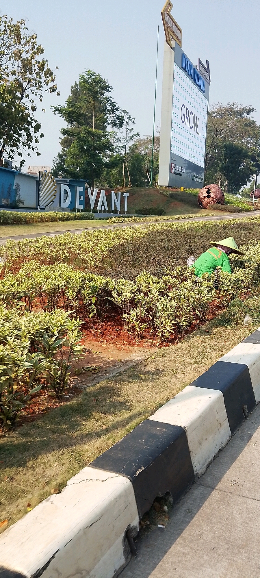 The image depicts a street scene in Gunung Putri, Indonesia, featuring a large sign for "KOTA WISATA" which displays the word "GROW...". To the left of this sign, another large sign reads "DEVANT". In the foreground, a person wearing a green shirt and a conical hat is tending to the bushes along the roadside. The environment includes well-maintained landscaping with small, leafy plants and red soil. A concrete barrier painted black and white runs along the edge of the road. Trees are visible in the background.