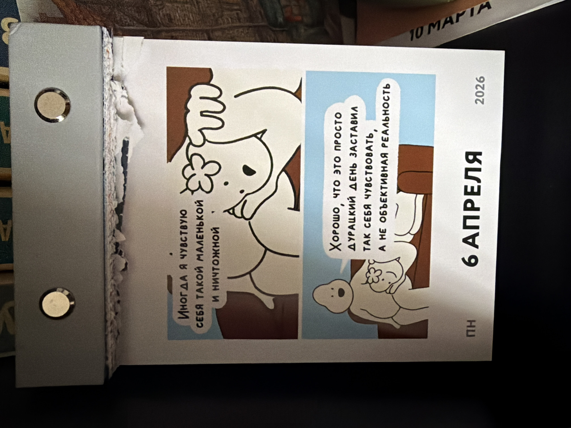 The image shows a page torn from a tear-off calendar, displaying the date April 6, 2026. The page features cartoon illustrations and text in Russian. The left side of the page is attached to a gray metal holder with two visible metal rivets. The edge of the calendar pages appears ragged and torn. The calendar is partially resting on a dark surface. Another calendar is also visible in the top right of the frame, showing "10 MAPTA" (March 10). There is no visible indication in the image showing that it is located in the city of Chaikovsky, Russia.