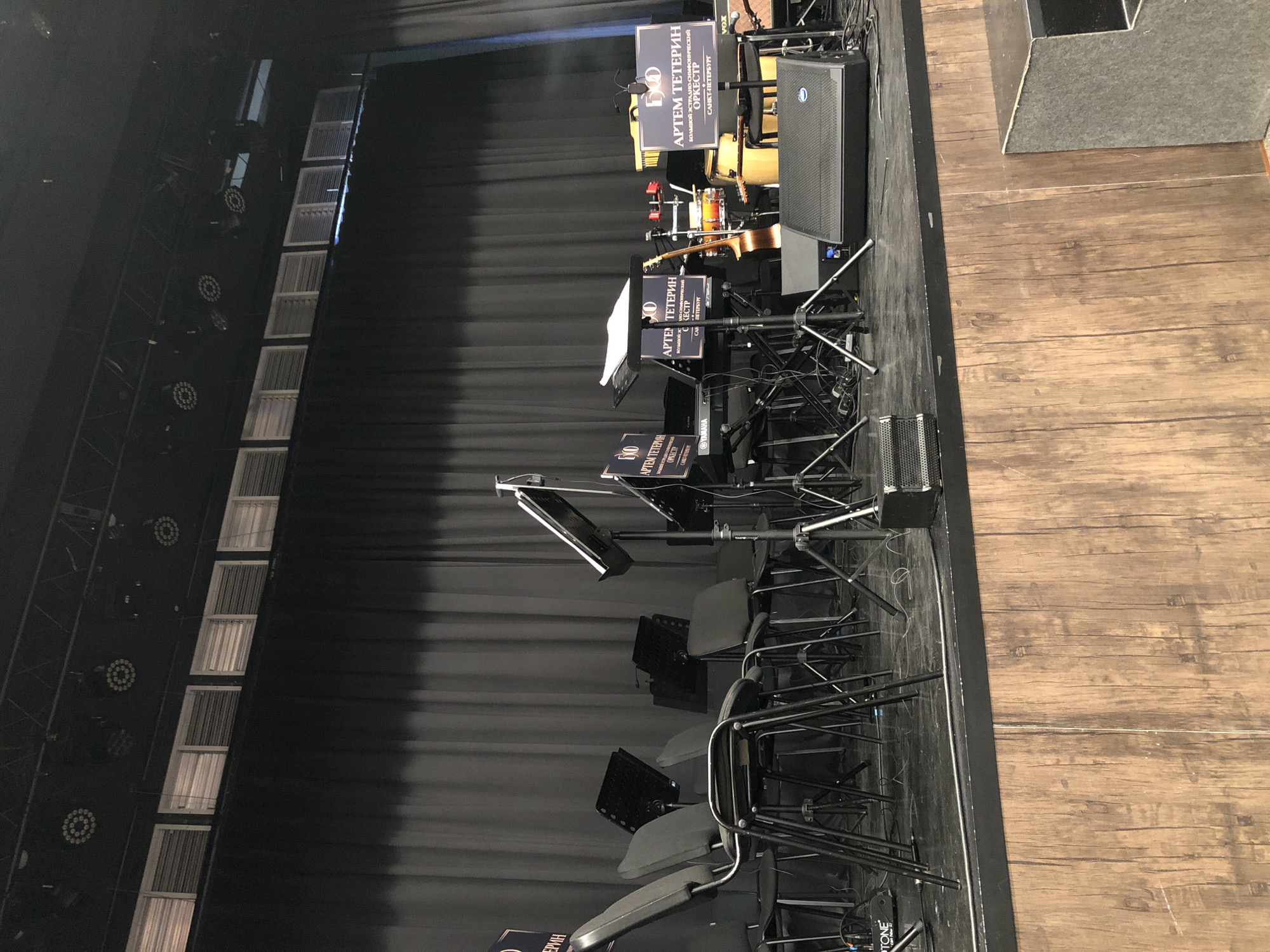 This is a photograph taken from above and to the side, looking down at a stage set up for a musical performance. The stage floor is made of weathered wooden planks. A black curtain serves as the backdrop, creating a theatrical atmosphere.
On the stage, several objects are arranged:
* **Musical Instruments:** An acoustic guitar is leaning against a drum set.
* **Audio Equipment:** A large black speaker is positioned on the right side of the stage. Several microphone stands are set up, some holding sheet music stands. A Yamaha keyboard is visible.
* **Seating:** A row of chairs, likely for the musicians or audience, is placed on the stage.
* **Signage:** Several rectangular signs with Cyrillic text are visible. One sign reads "АРТЕМ ТЕТЕРИН БОЛЬШОЙ ЭСТРАДНО-СИМФОНИЧЕСКИЙ ОРКЕСТР САНКТ-ПЕТЕРБУРГ" which translates to "Artem Teterin Grand Estrada-Symphonic Orchestra Saint Petersburg". Another sign says "VOX".
The scene appears to be indoors, possibly a concert hall or theater, given the stage setup and lighting rigs visible in the upper left corner. There are no people visible in the photograph. The overall impression is one of preparation for a performance, with the stage ready for musicians to arrive. The lighting suggests it might be daytime, but the interior setting makes it difficult to determine definitively. The context provided indicates the location is Moscow, Russia.