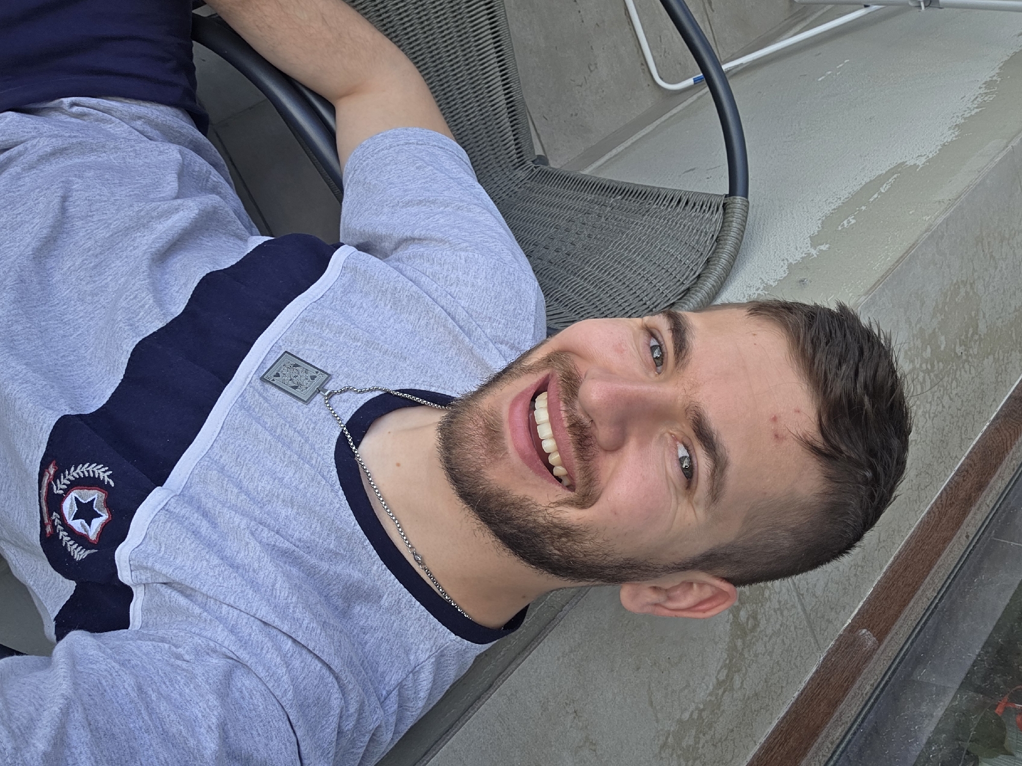 A young man with short brown hair and a beard is smiling while sitting in a wicker chair on a tiled balcony. He is wearing a grey t-shirt with a navy blue stripe on the shoulder. A silver chain with a pendant is visible around his neck. The location is inferred to be Yagodovo, Bulgaria, based on the user prompt. A white electrical conduit runs along the wall in the background. Part of a window or glass door is visible to the right.