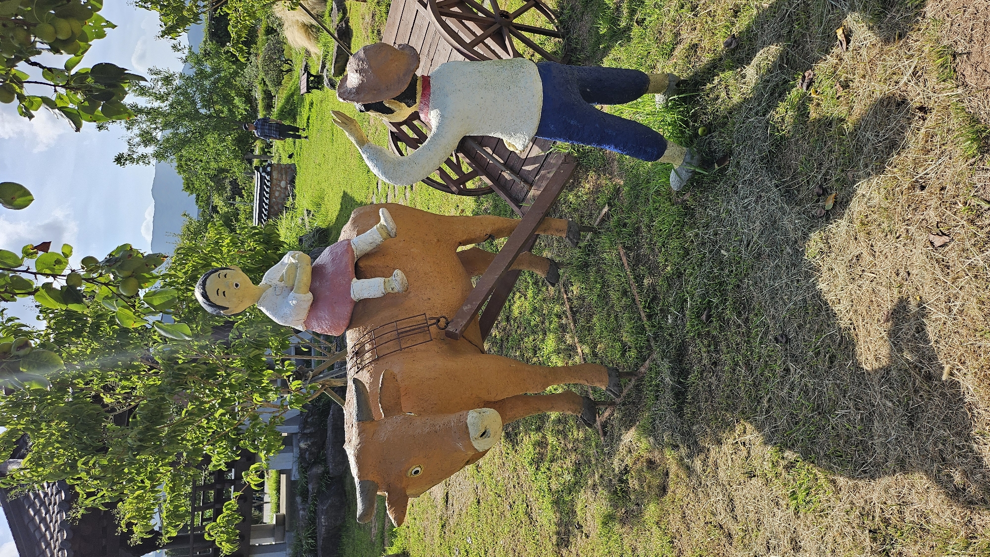 This outdoor image depicts a scenic view with sculptures in the foreground and a natural landscape in the background. The sculptures represent a farmer guiding a bull, with a child seated on the bull. The farmer, dressed in a white shirt and blue pants, is facing away from the viewer, with his right arm raised as if to direct or communicate. The bull, painted in an earthy brown hue, is harnessed to a wooden cart, with the child, dressed in a pink skirt and white top, seated atop the bull. The scene is set in a grassy area with some dry patches and fallen leaves, suggesting a park or rural environment. In the background, there is a traditional Korean tiled roof structure, lush green trees, and mountains in the distance under a clear blue sky with scattered clouds. A person in casual attire can be seen walking in the middle ground. This scene is located in Geoje-si, South Korea, likely at a cultural or historical attraction that showcases traditional Korean life.