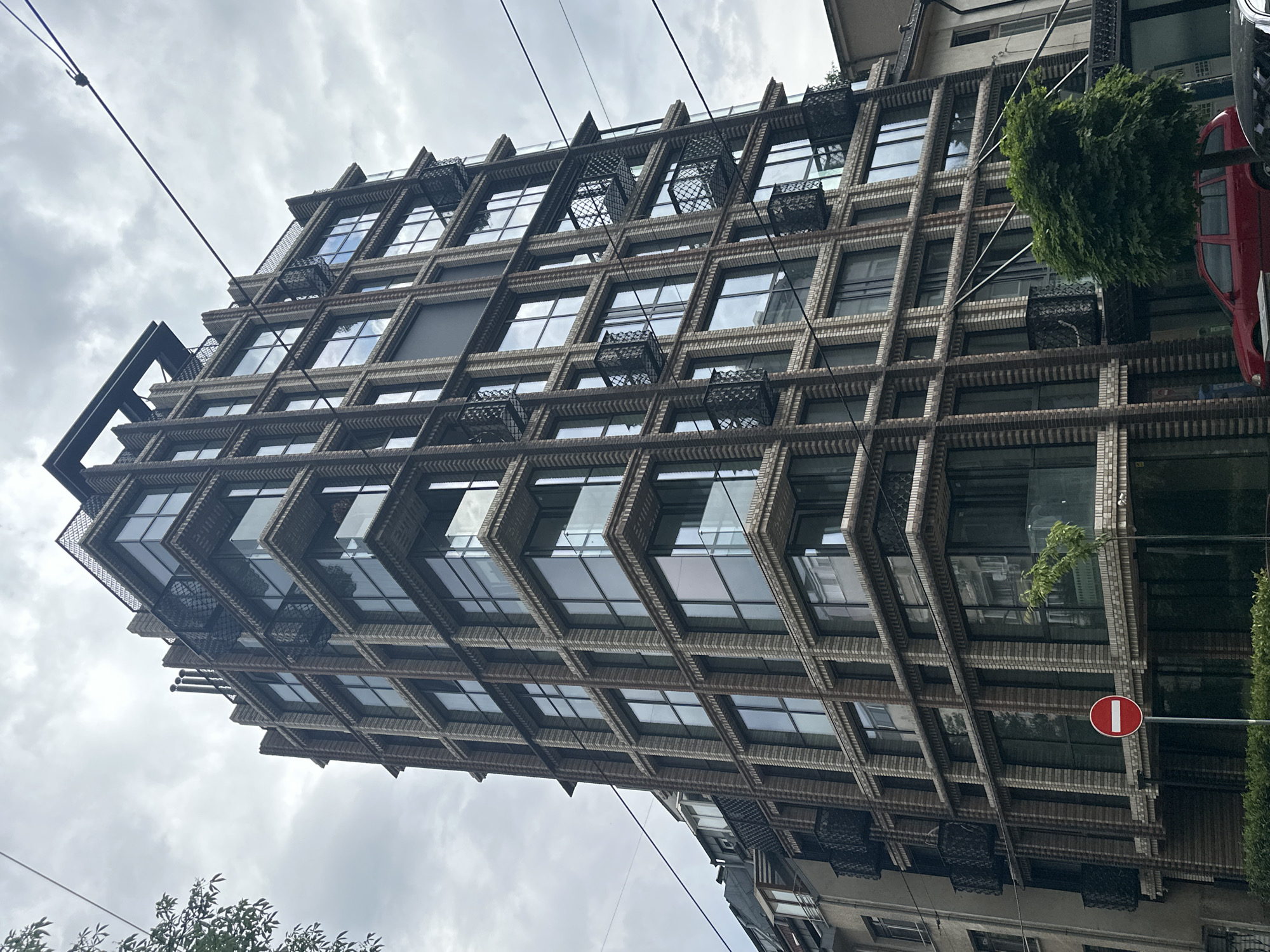 The image shows a multi-story building in Sofia, Bulgaria, viewed from a low angle. The building has a grid-like facade with rows of windows recessed within a brick or stone framework. Some windows have decorative ironwork or planters. The sky is overcast. To the right of the building, there is a red vehicle (possibly a car or van), a green tree, and a red and white "do not enter" traffic sign. Power lines are visible against the sky. The building exhibits modern architectural design. Other buildings are partially visible at the bottom and top right of the image.