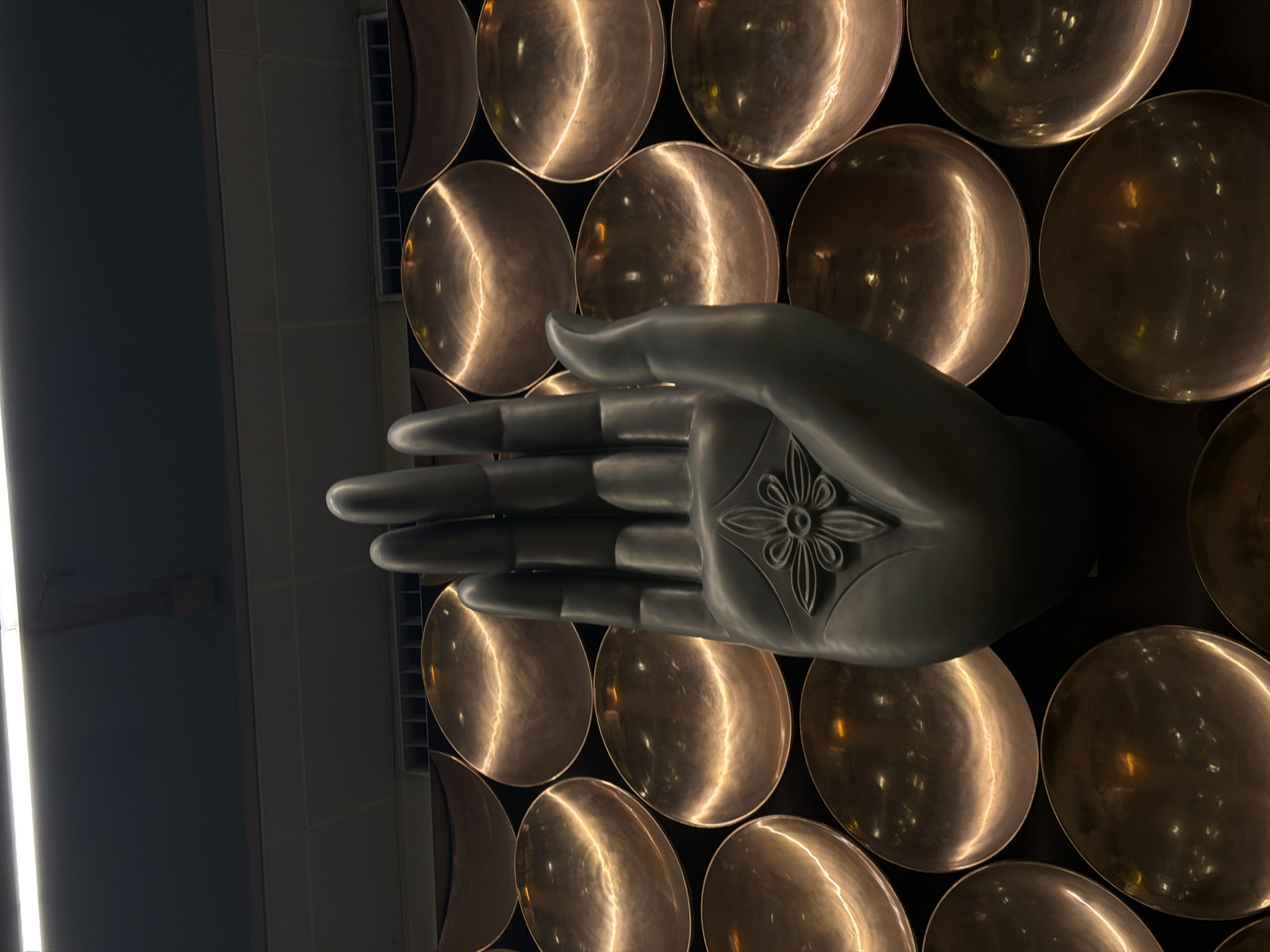 The image shows a close-up of a large, dark gray, stylized hand sculpture with its palm facing forward, adorned with a floral motif. The hand is positioned in front of a patterned background composed of many round, reflective metallic dishes, likely copper or brass, with a warm, golden sheen. These dishes are arranged in a honeycomb-like pattern, creating a visually rich texture. The lighting is dramatic, with strong highlights on the metallic dishes and a softer, diffused light on the sculpture. The top of the image reveals a dark ceiling with a fluorescent light fixture, suggesting an indoor or covered space. This sculpture and its backdrop are located in Terminal 3 of the Indira Gandhi International Airport in Delhi, India, where it serves as an art installation.