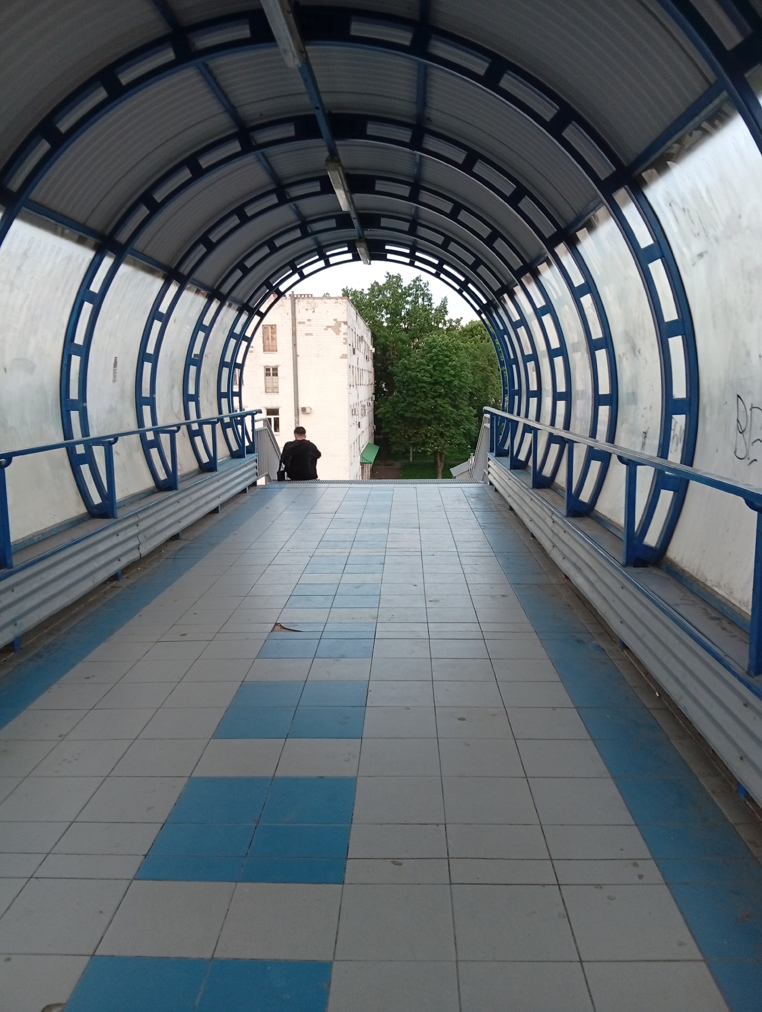 A full shot depicts a pedestrian overpass in Poltava, Ukraine. The overpass has a curved, blue and white metal frame with a corrugated metal roof. Blue handrails line the sides of the walkway. The walkway floor is paved with alternating grey and blue square tiles.
A lone man, dressed in black, is seated at the far end of the overpass, near the stairs leading down. Beyond the overpass exit, a white building and lush green trees are visible.