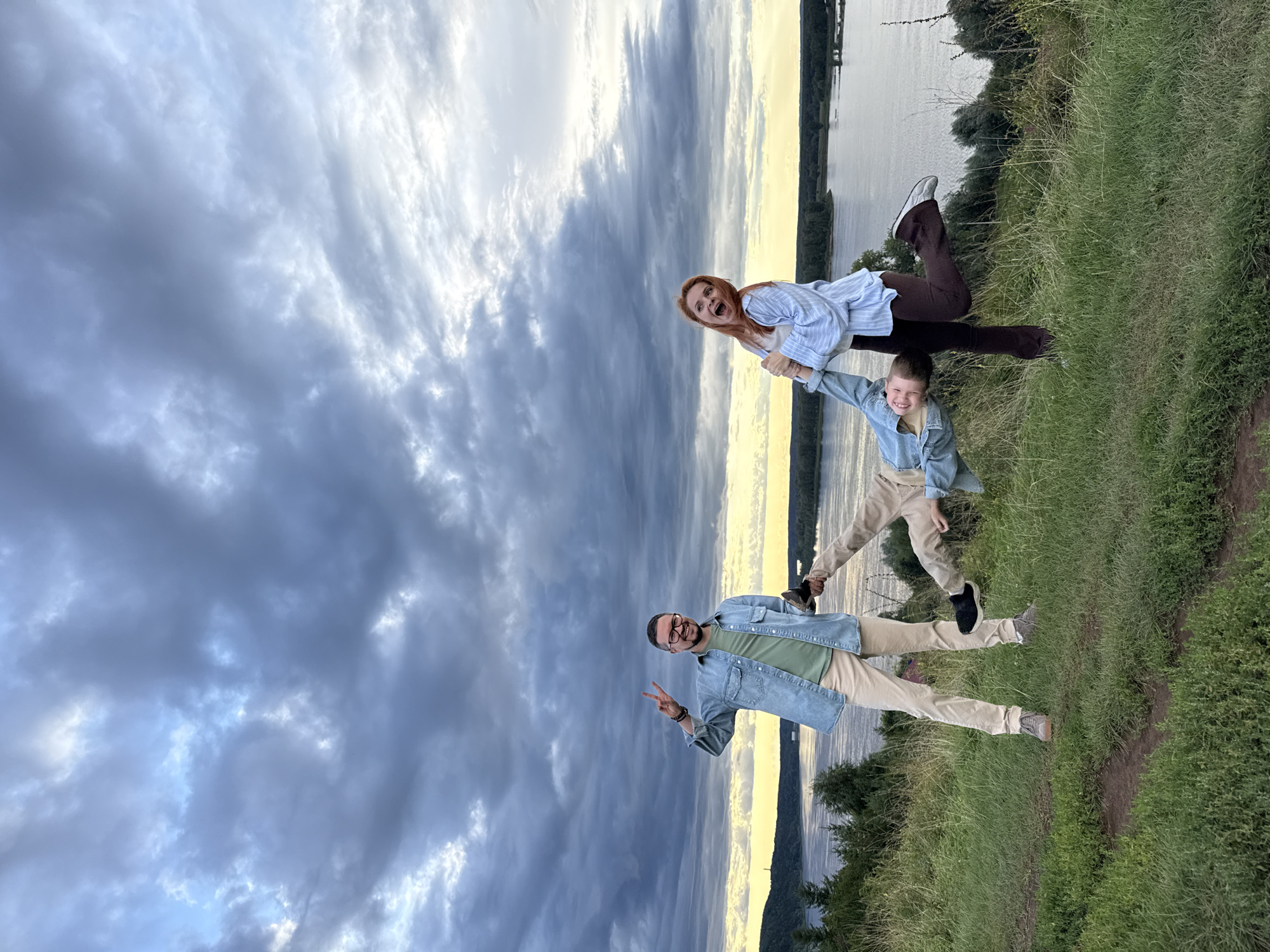 Three people are on a grassy hill overlooking a body of water. A man in a denim shirt and beige pants is on the left, smiling and holding up two fingers. In the center, a person in a denim jacket and beige pants is being held upside down by their legs by a woman on the right. The woman has red hair, is wearing a blue and white striped shirt and brown pants, and is laughing with her mouth open. The sky is cloudy with patches of blue and yellow light. The body of water is wide and calm, with a tree-lined shore on the opposite side. The location appears to be the city of Olkhovka, Russia, given the description of the landscape which matches typical views of the riverfront there. The overall atmosphere is playful and joyful.