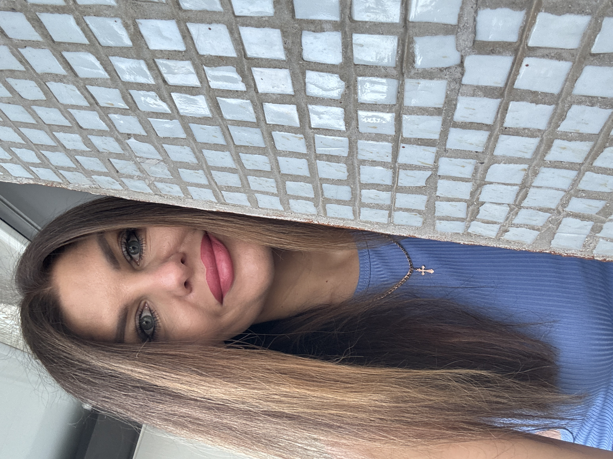 A woman with long, brown, wavy hair and blue eyes is partially obscured by a wall covered in small, light blue square tiles. She is wearing a blue ribbed tank top and a delicate gold necklace with a cross pendant. Her lips are painted a bright pink. The background shows a hint of a window or door frame. The location is the city of Chaikovsky, Russia.