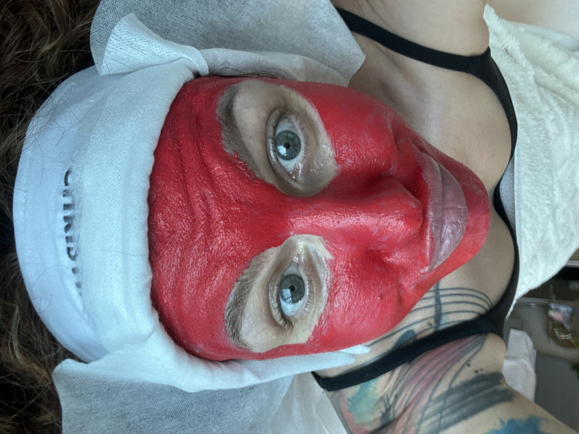 A person lies supine with a red facial mask applied. The mask covers the entire face except the eyes. A white fabric head covering is also present. The person appears to be female, wearing a black garment and having tattoos on their chest and arm. A white towel is visible in the background. The city is not discernible in this image.
