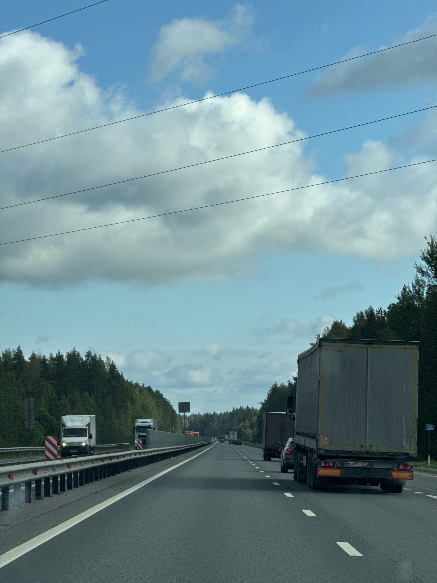 The image captures a view from a vehicle traveling on a highway, with a dense forest lining the right side of the road. Several large trucks are visible, indicating commercial traffic. One truck, in the foreground, has a license plate that reads "EA 8509 77". The sky is partly cloudy, with large, white clouds against a blue background. Power lines are strung across the upper portion of the image. The highway appears to be in a rural or semi-rural setting, likely outside a major urban center. The location is identified as Krasnye Stanki, Russia.