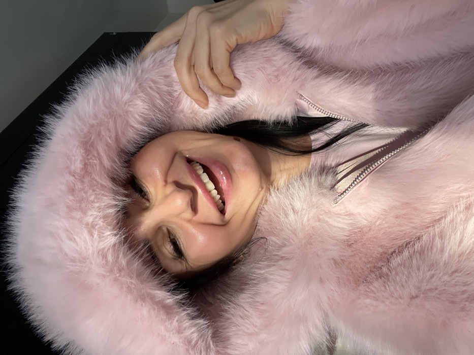 An adult female is the sole subject of a close-up portrait taken in Syasstroy, Russia. She has dark hair and a visible mole on her lower left jawline. The woman is smiling broadly, revealing her upper and lower teeth, with her lips appearing glossy and pink. She is wearing a light pink, thick, furry hooded jacket or coat, with a prominent silver-colored zipper visible on the front. A lighter pink garment is visible underneath the jacket. Her right hand is raised, with fingers slightly curled, lightly touching the fur of the hood near her right cheek. The background is dark and out of focus, with an indistinct dark rectangular shape present in the upper right quadrant, suggesting an indoor setting or a closely proximate dark structure.