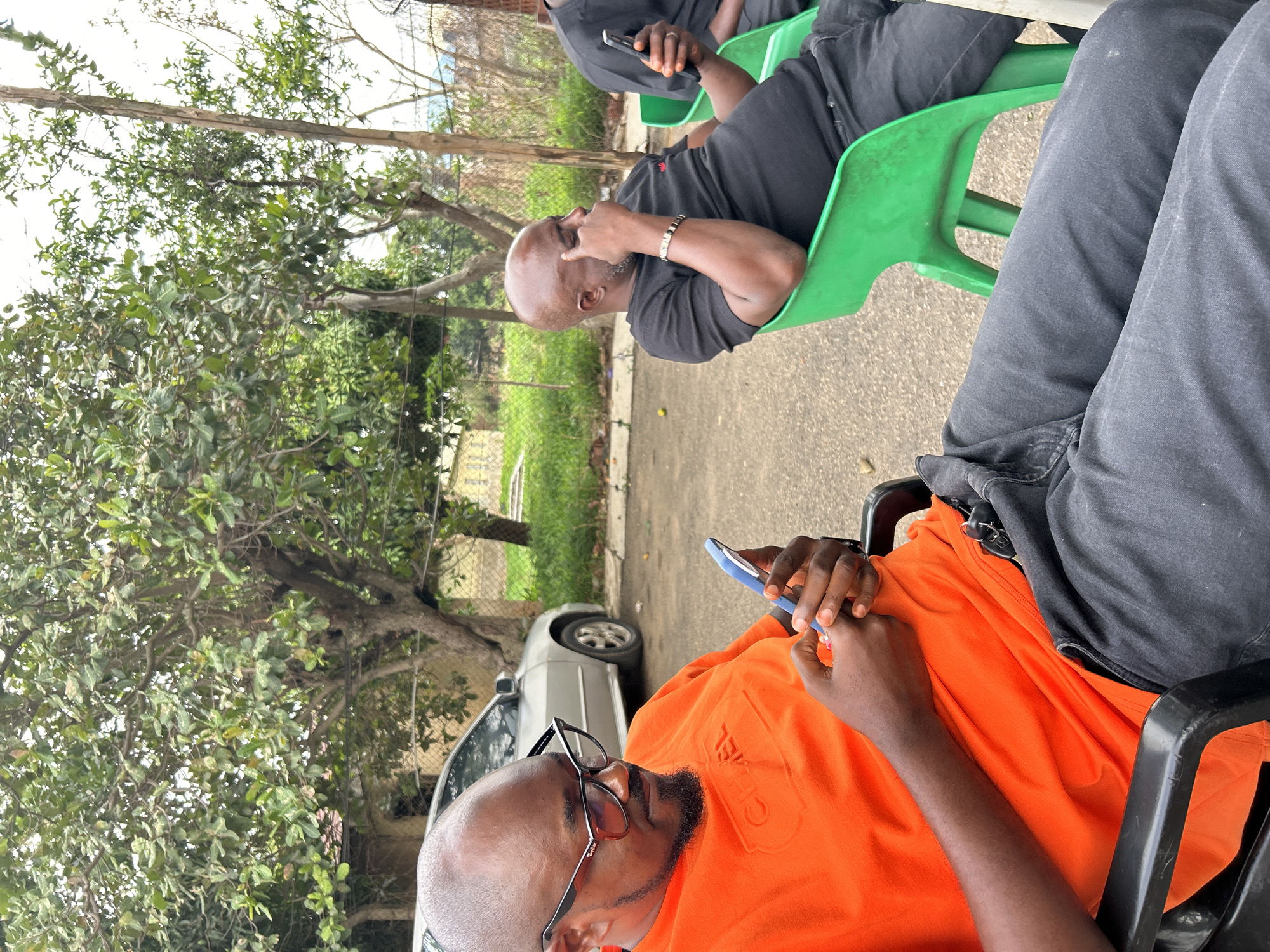 Several African men are seated outdoors in Jos, Nigeria. The man in the foreground, wearing an orange shirt and glasses, is focused on a cell phone. To the left, a parked silver car is visible near trees. Further back, another bald man in a black shirt is seated in a green plastic chair, with another person in the background looking at a cell phone.