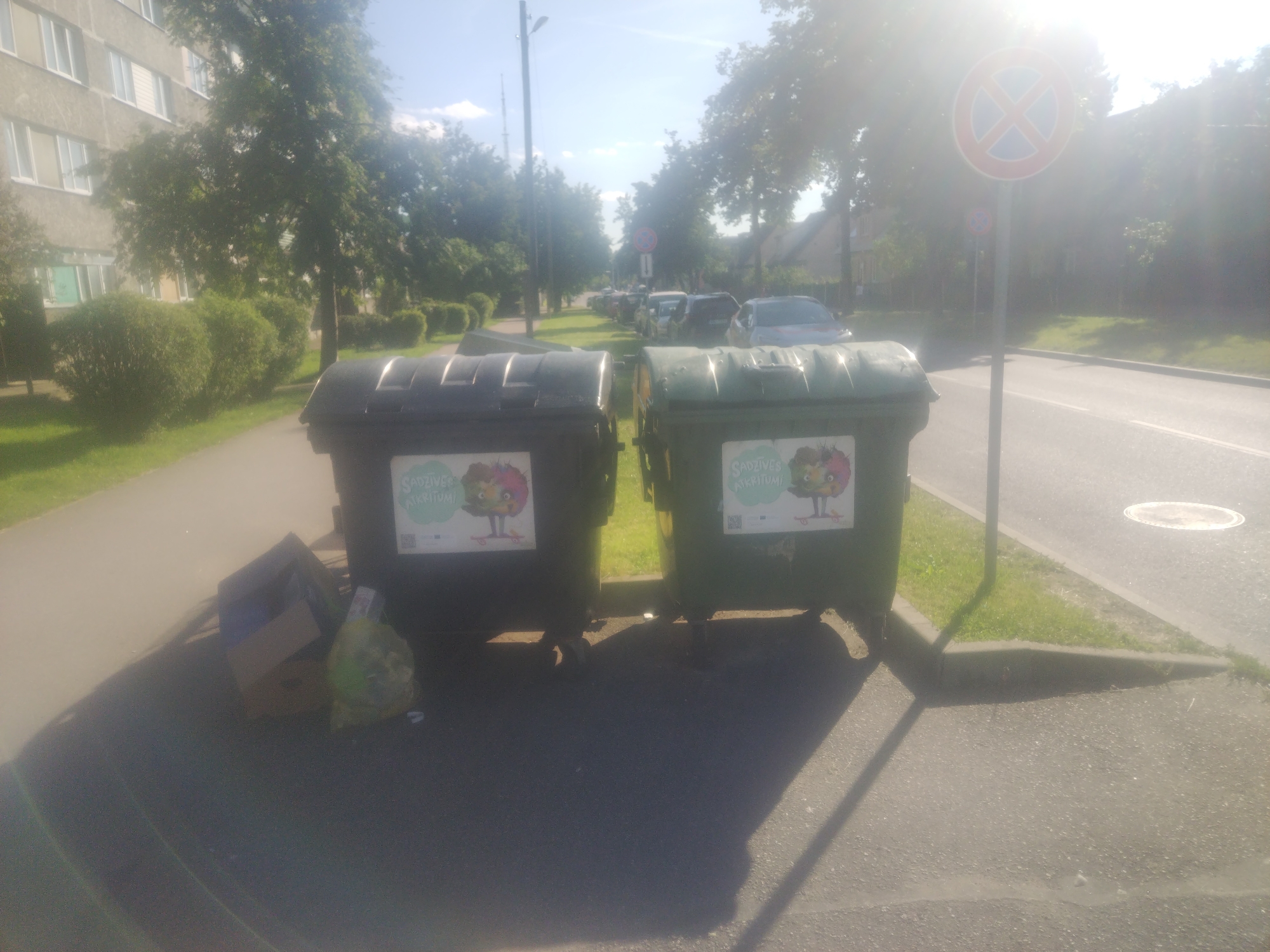 The image depicts an urban scene in Daugavpils, Latvia, on a sunny day. In the foreground, two large green and black waste bins stand on a paved area adjacent to a sidewalk. Both bins have decals featuring cartoon characters and text in a foreign language, possibly Latvian. To the left of the bins, a flattened cardboard box and a plastic bag filled with what appears to be trash are visible.
The background shows a tree-lined street with parked cars. A tall, thin lamppost is positioned behind the bins. Further down the street, a traffic sign with a red circle and a blue cross inside, indicating no parking or stopping, is mounted on a pole. Beyond the street, buildings and more trees are discernible. The overall environment suggests a residential or commercial area within the city. The lighting indicates it is daytime, with the sun casting shadows.