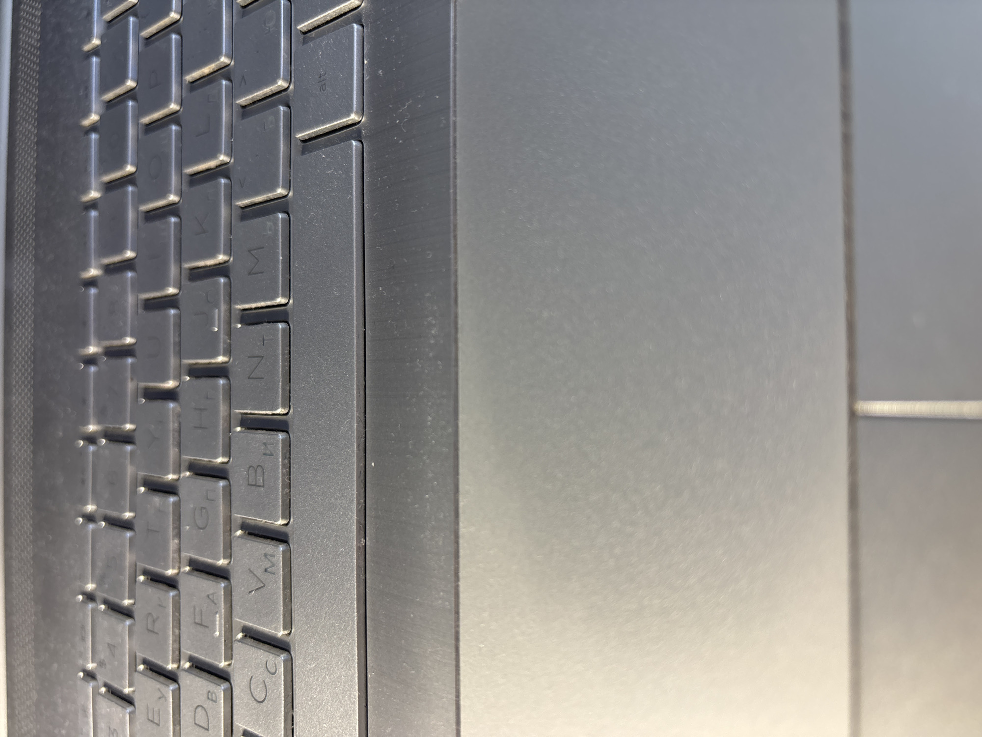 This is a close-up, top-down, medium shot of a laptop keyboard. The keyboard is made of dark gray plastic with individual keys. The keys visible include letters and symbols, with some keys showing Cyrillic characters, suggesting a Ukrainian or Russian keyboard layout. Some keys visible are 'E', 'R', 'T', 'Y', 'D', 'B', 'F', 'G', 'H', 'C', 'V', 'B', 'N', and 'M'. The 'Alt' key is also visible. The keyboard is slightly dusty. The area below the keyboard is a smooth, light gray surface, possibly the palm rest of the laptop. The lighting is bright and appears to be coming from the left, casting shadows on the right side of the keys. The overall environment is indoors, likely a desk or table. There are no people, animals, or specific landmarks visible that would indicate a location within the city of Bila Tserkva, Ukraine.