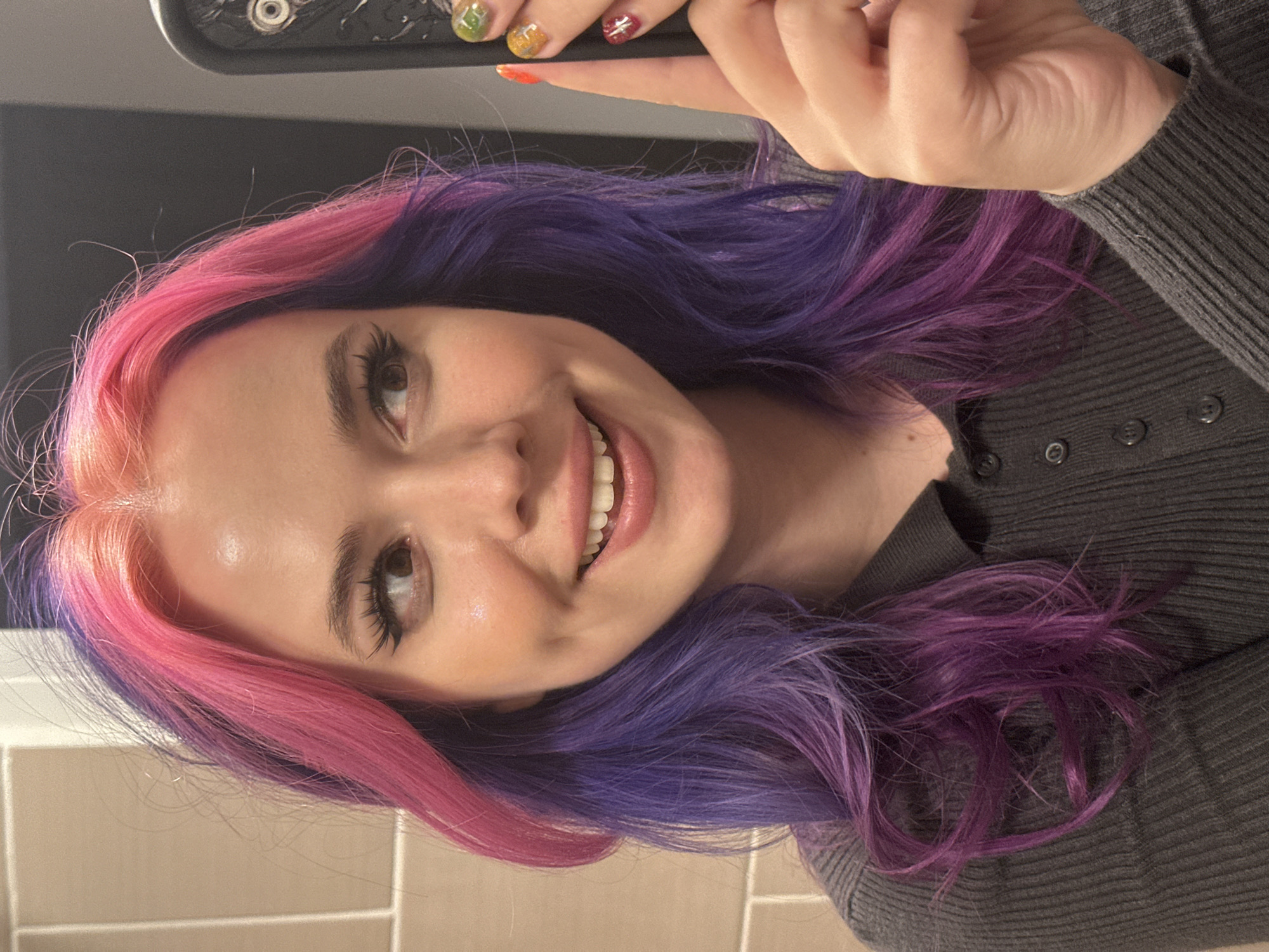 A person with pink and purple hair is facing the camera in an indoor setting, possibly a bathroom with beige tiles visible in the lower left corner. The person is smiling and holding up a phone, suggesting they are taking a selfie. They are wearing a gray, button-down sweater. The person's nails have different colors. There is no clear indication of City of Zagreb, Croatia, within this image.