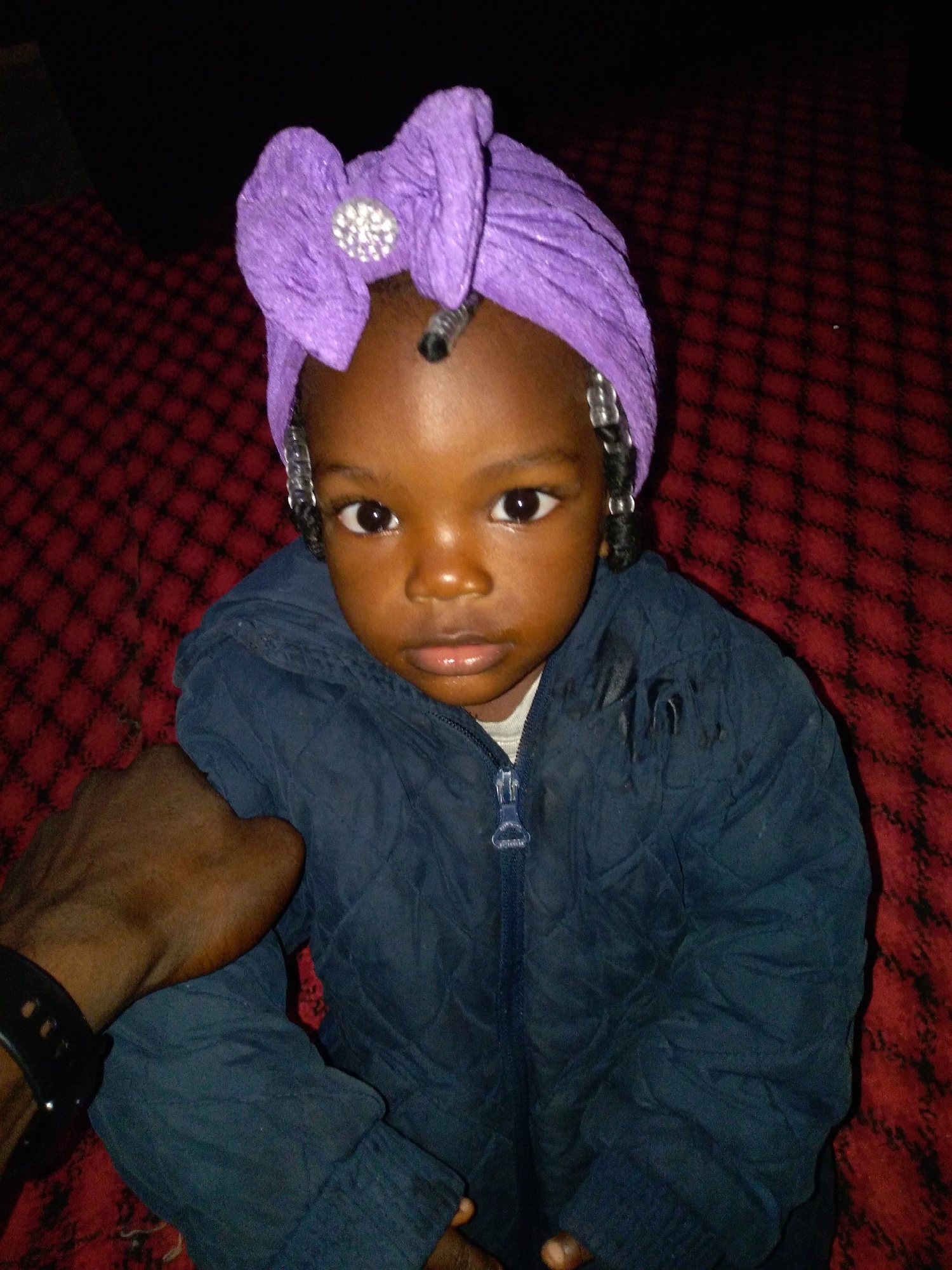 A young Black child, likely a toddler, is the central subject of the photograph, seated on a red and black patterned surface. The child is wearing a dark blue quilted jacket with a zipper closure, and a vibrant purple headwrap adorned with a sparkly, silver, flower-shaped embellishment. The child's hair is styled in braids with silver beads. The child's left arm is resting on their lap, and their right hand is visible, appearing to be held by an adult. The background is dark, suggesting an indoor setting. The lighting appears to be artificial, casting shadows and highlighting the child's face. The overall impression is of a personal, informal portrait.