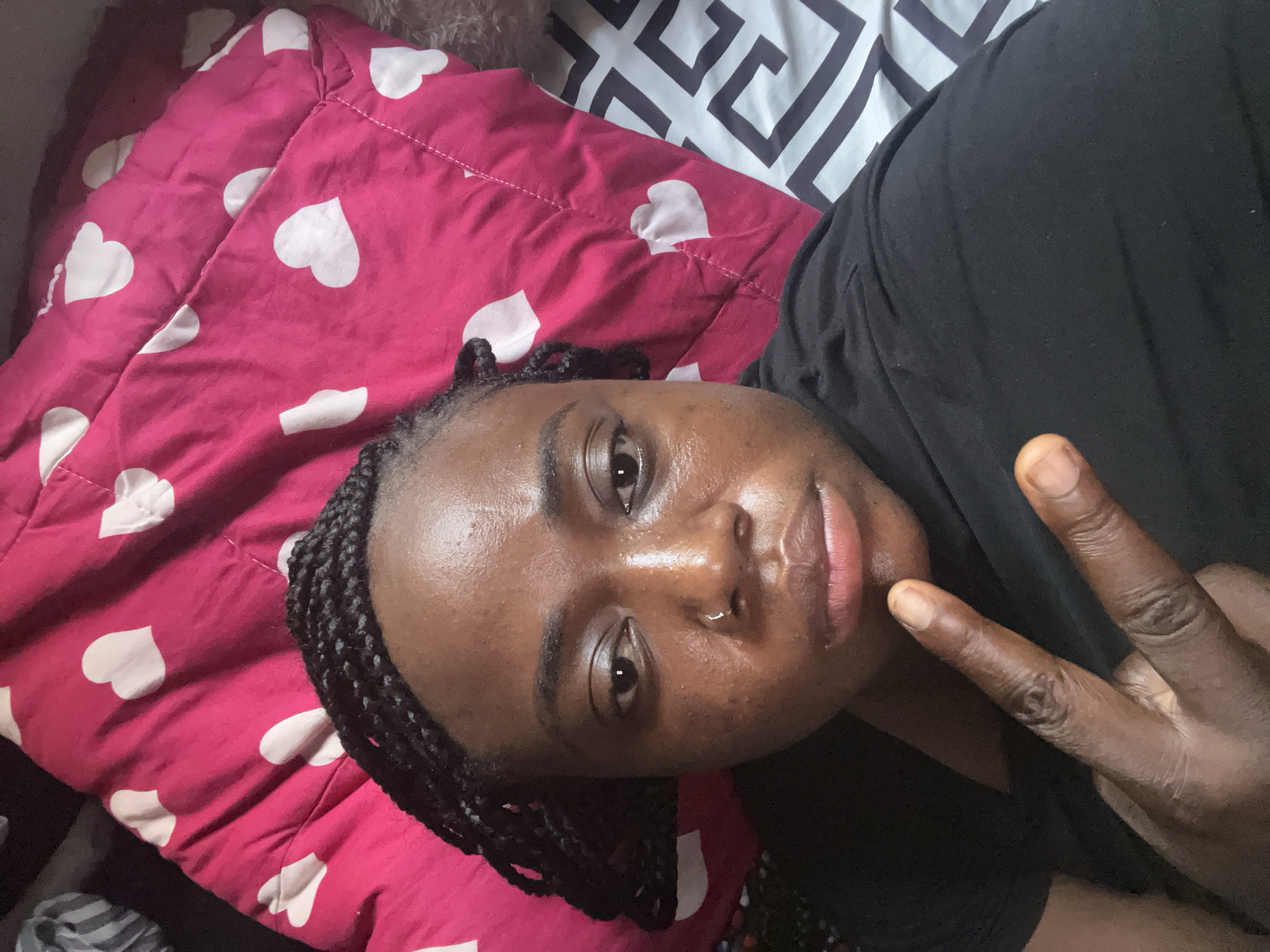 The image is a close-up, angled selfie of a dark-skinned individual lying down, making a peace sign with their right hand. The person has braided hair pulled back, a subtle silver nose ring in their left nostril, and is wearing a black t-shirt. Their expression is neutral, and their skin shows a natural texture with some minor blemishes. They are resting on a vibrant pink duvet or comforter decorated with numerous white heart patterns. Part of a second piece of bedding with a white and dark geometric design is visible in the upper background. The indoor lighting is soft and ambient. No text is visible in the image. This scene takes place in Abuja, Nigeria.